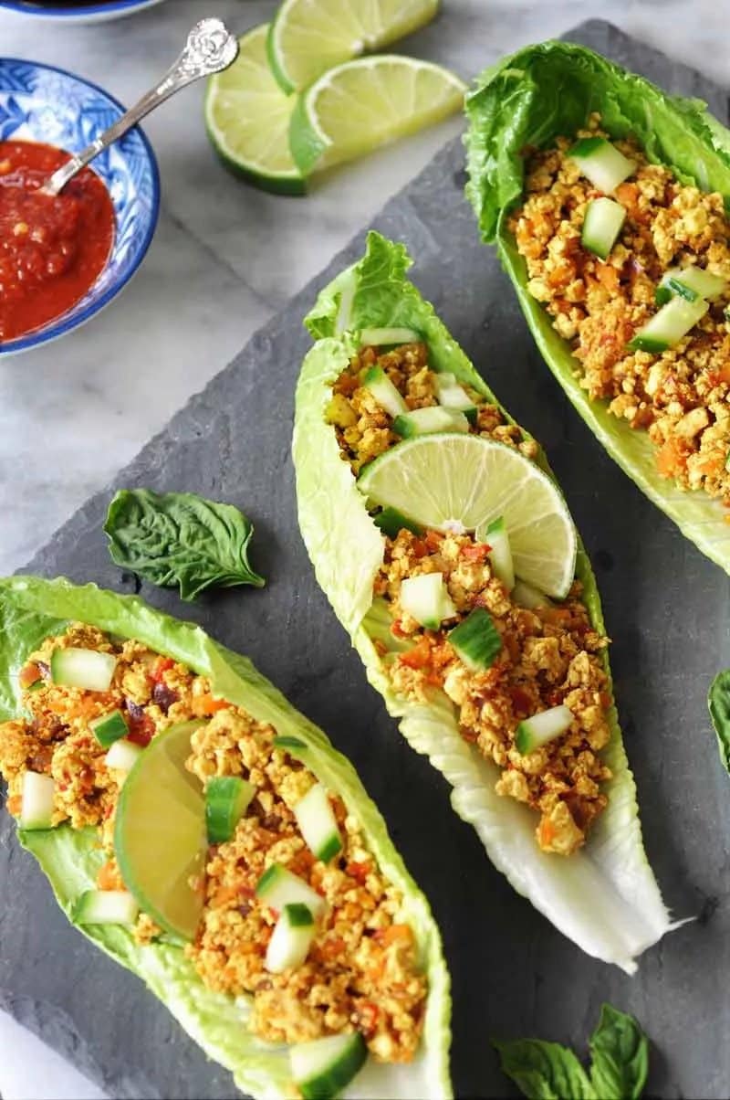Three tofu lettuace wraps with a chopped lime on top situated on a black charcoal chopping board. In the corner of the photo you see a small bowl of red dipping sauce.