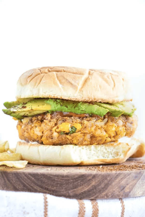 Chickpea pattie with avocado slices served on a wooden board with a side of crisps.