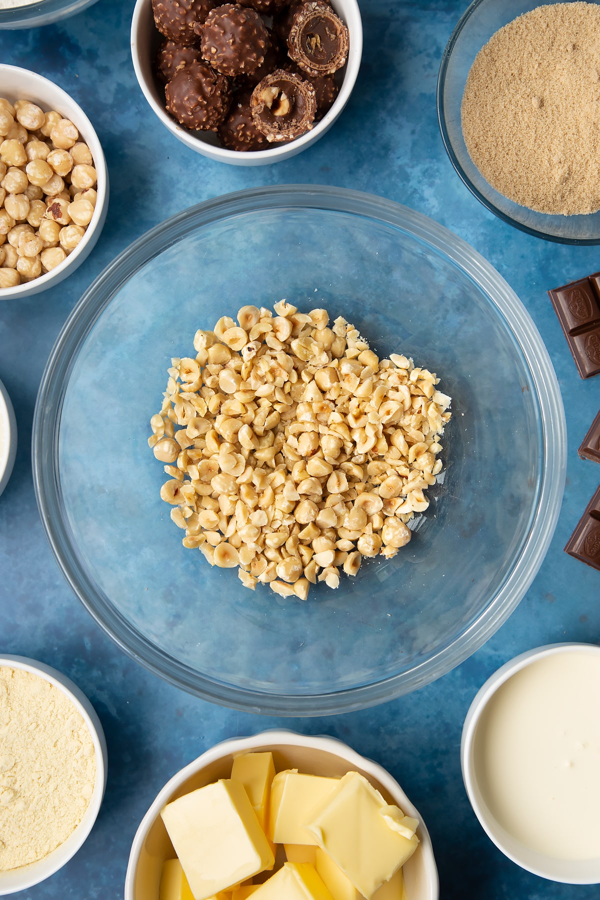 Chopped hazelnuts in a glass mixing bowl. Ingredients to make a Ferrero Rocher slice surround the bowl.