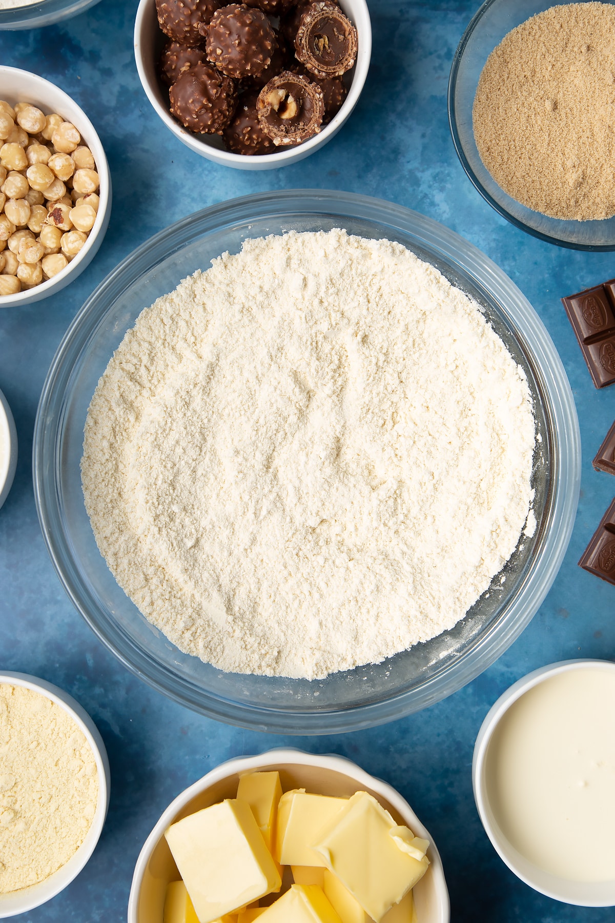 Flour, sugar and semolina mixed together in a glass mixing bowl. Ingredients to make a Ferrero Rocher slice surround the bowl.