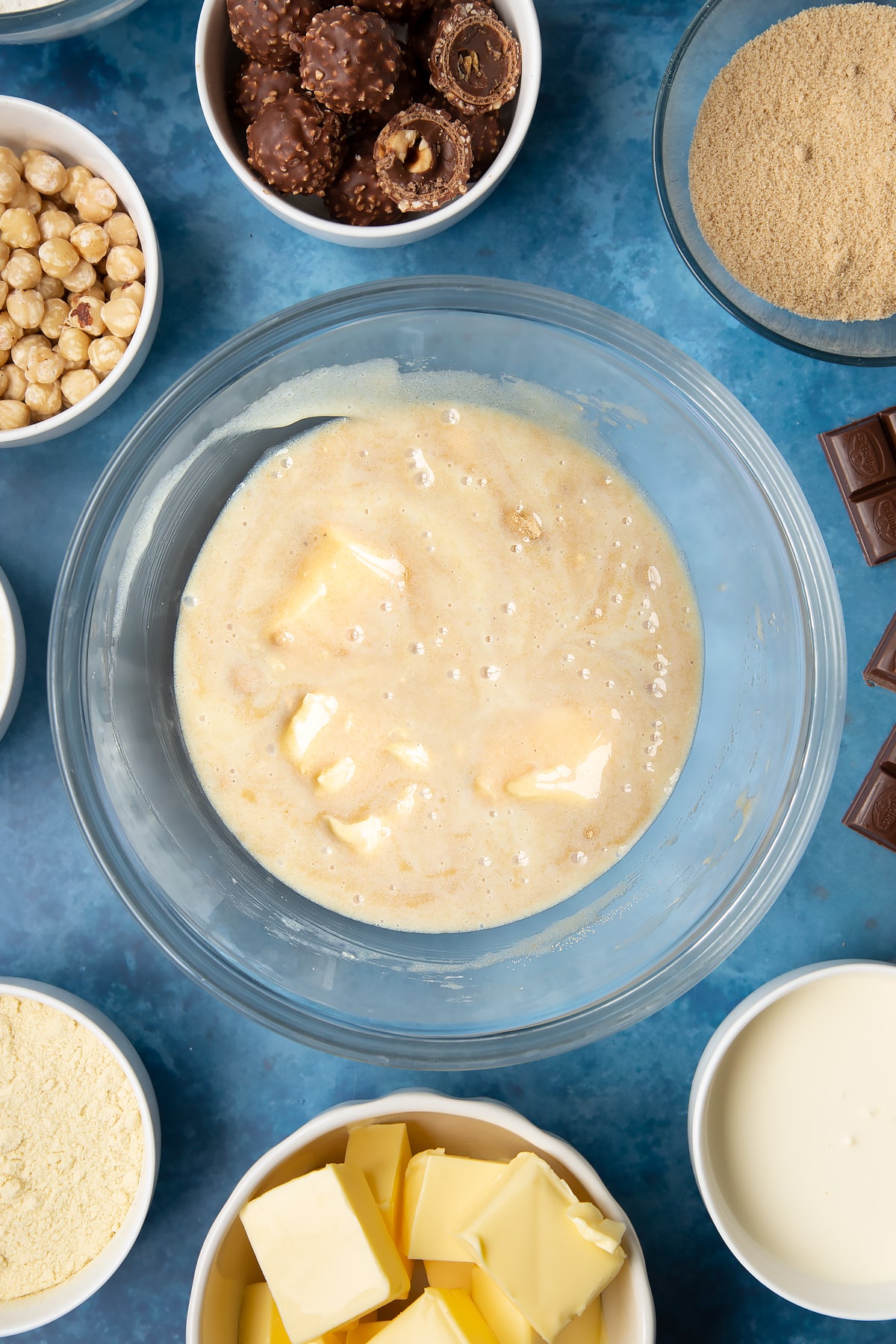 Brown sugar, butter and condensed milk stirred together in a glass mixing bowl. Ingredients to make a Ferrero Rocher slice surround the bowl.
