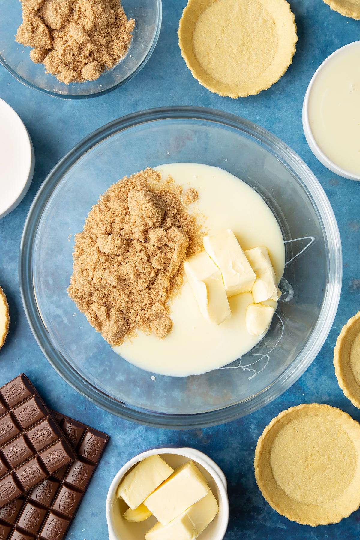 Light brown sugar, butter and condensed milk in a large mixing bowl. Ingredients to make mini chocolate tarts surround the bowl.