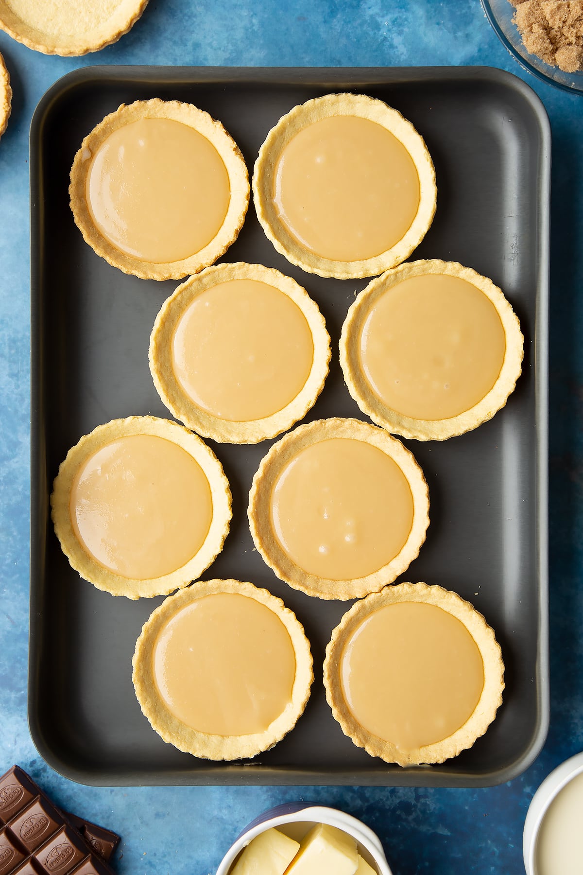 Small pastry cases filled with caramel on a large metal baking tray. Ingredients to make mini chocolate tarts surround the tray.
