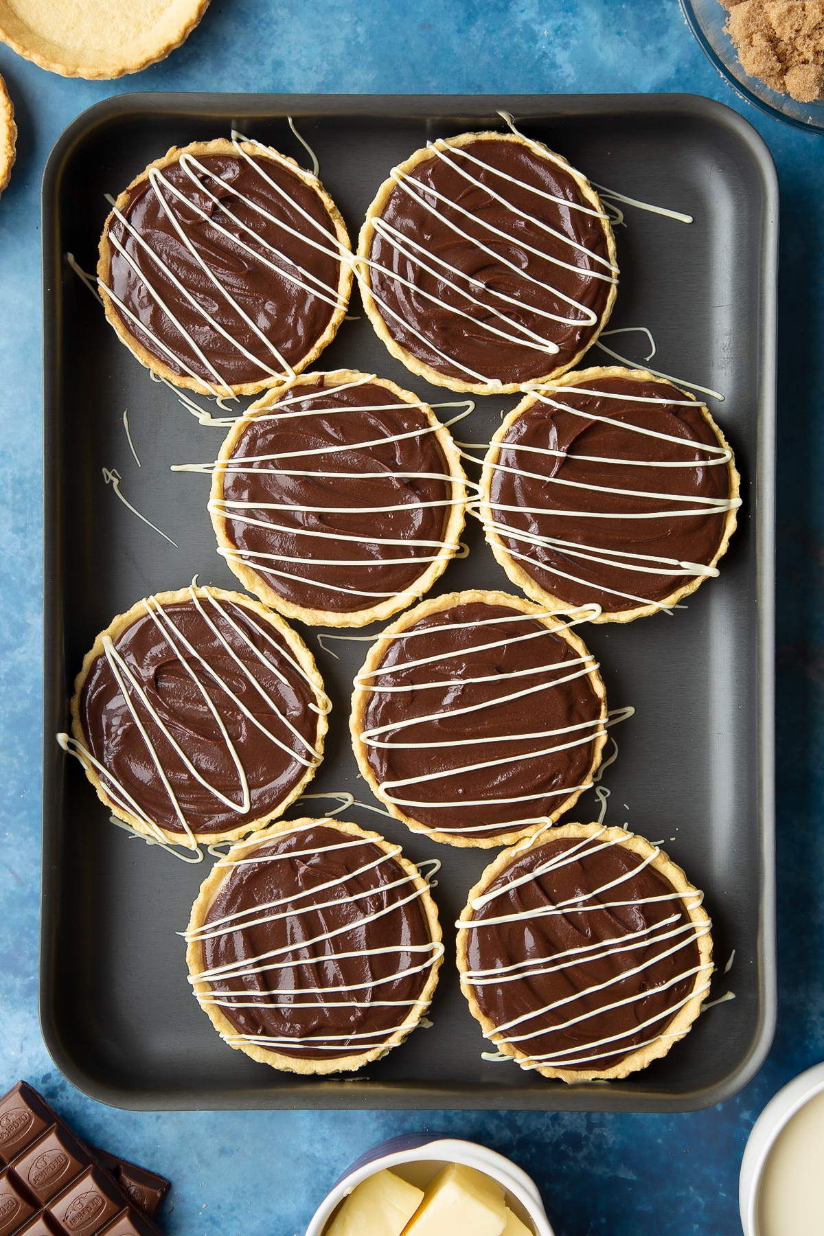 Mini chocolate tarts decorated with drizzled white chocolate on a large metal tray.