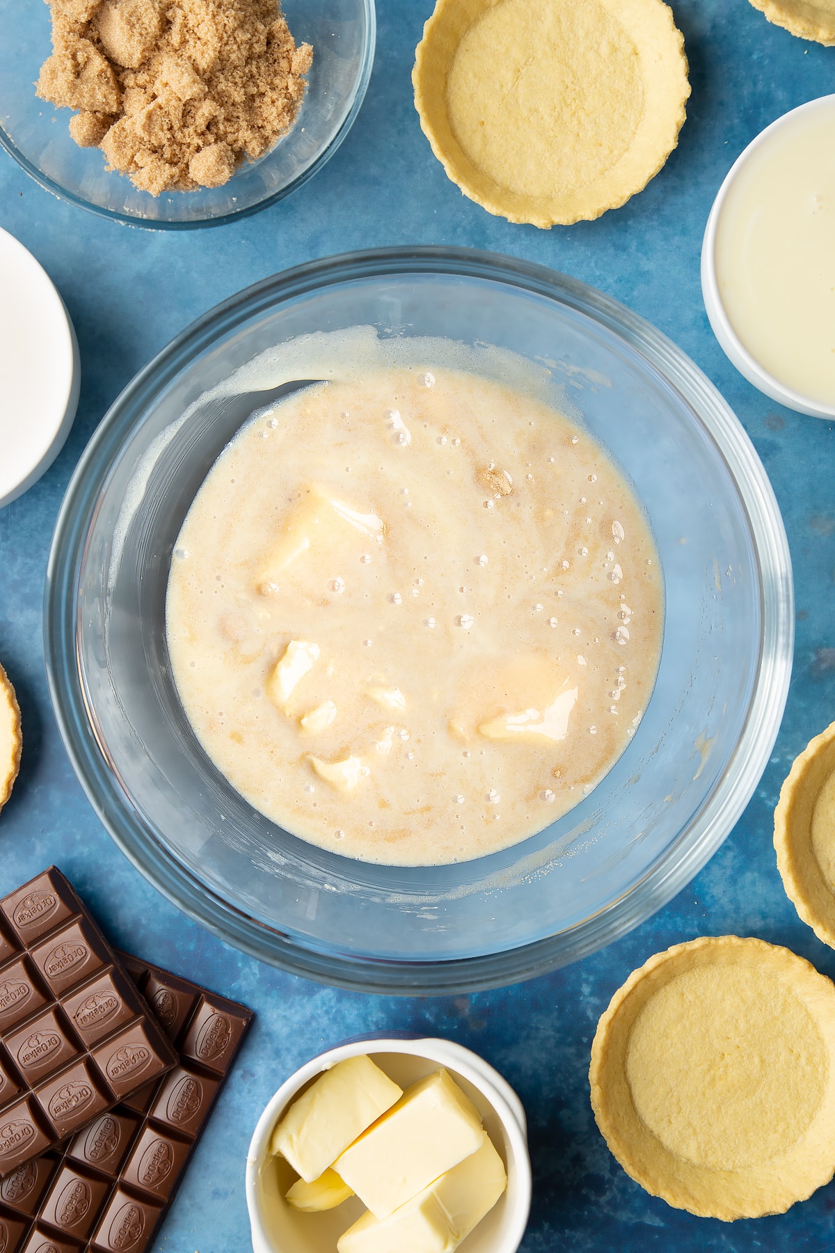 Light brown sugar, butter and condensed milk stirred together in a large mixing bowl. Ingredients to make mini chocolate tarts surround the bowl.