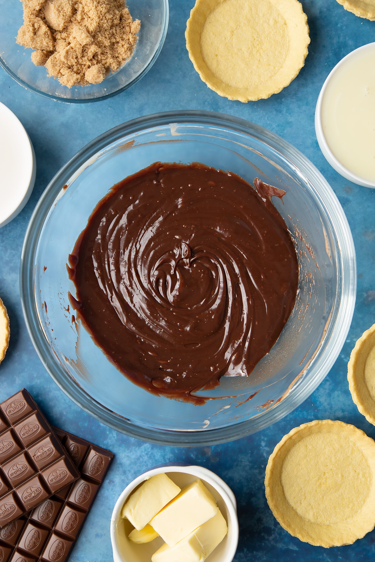 Freshly made milk chocolate ganache in a large mixing bowl. Ingredients to make mini chocolate tarts surround the bowl.