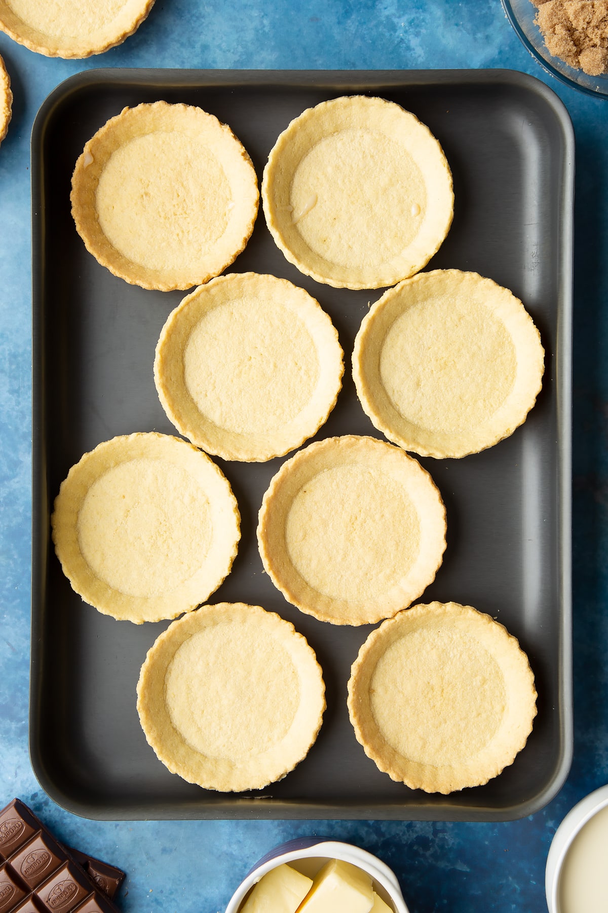 Small pastry cases on a large metal baking tray. Ingredients to make mini chocolate tarts surround the tray.