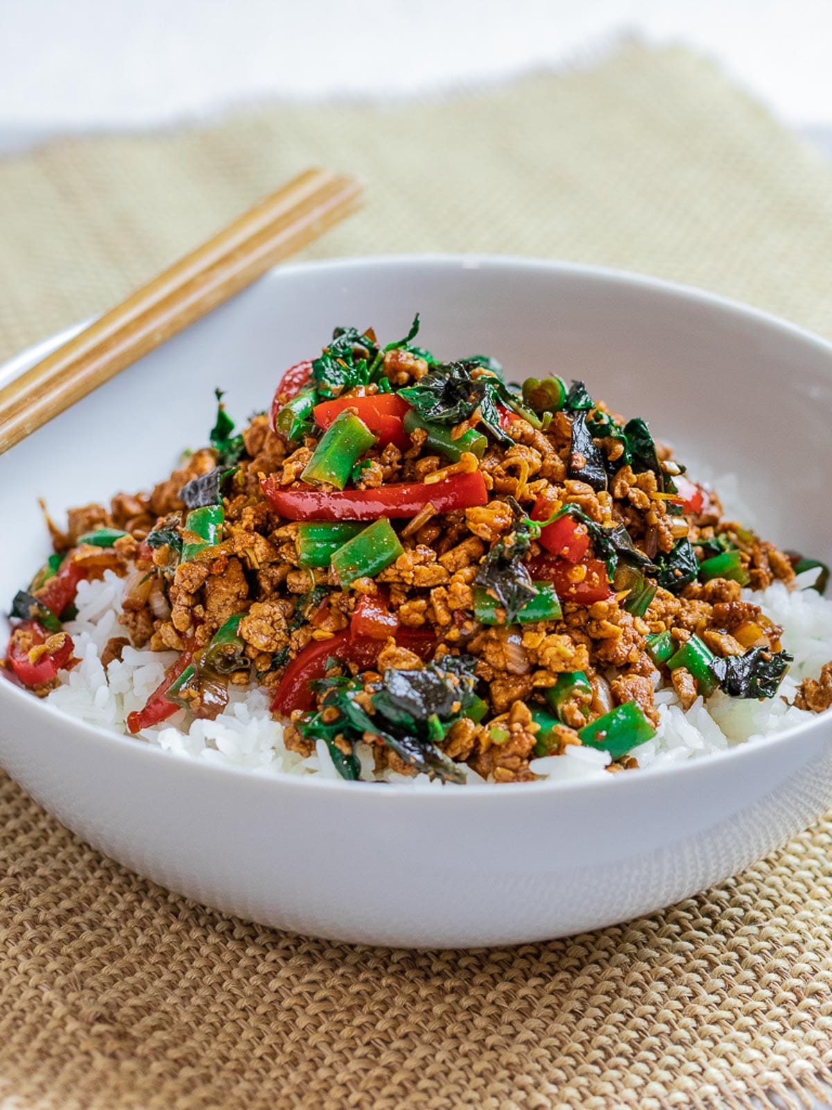A bowl of Thai basil tofu stir fry, also known as pad krapow sits on an organic table cloth with chopsticks on the side.
