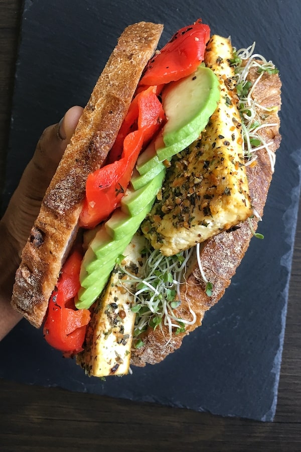 Crusty bread slices stacked between tofu, avocado and roasted red peppers held together with a hand.