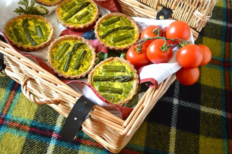 Five mini Asparagus Tarts With A Pesto Surprise sit inside a wicker basket. At the side are some on the vine cherry tomatoes. The basket sits on a deocrative checked rug.