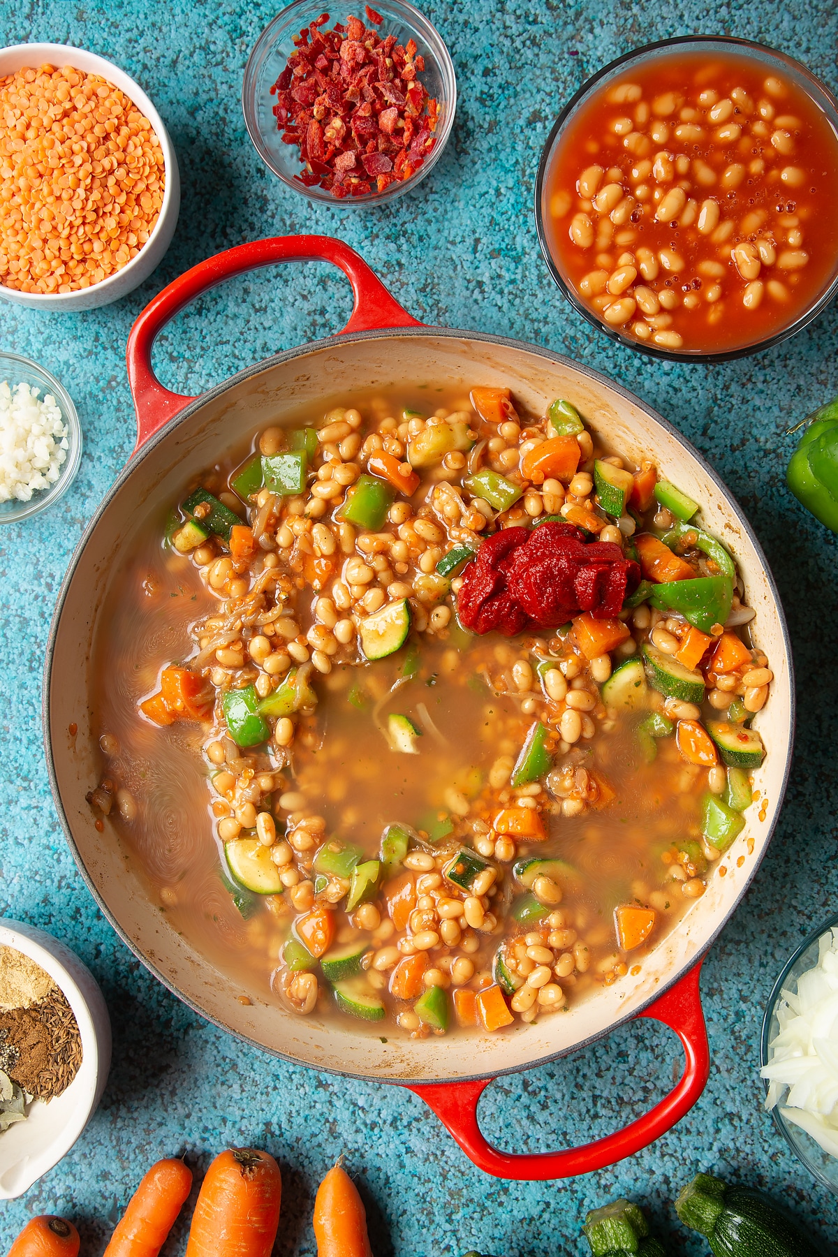 Fried onions, spices, peppers, carrots, courgette, red lentils and baked beans in a pan. Stock and tomato puree sit on top. Ingredients to make baked bean curry surround the pen.