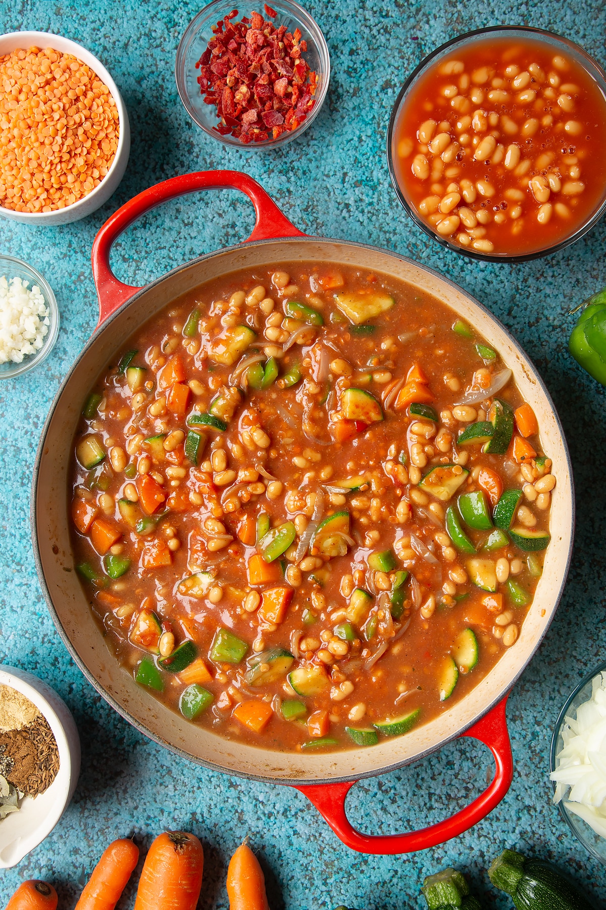 Fried onions, spices, peppers, carrots, courgette, red lentils, baked beans stock and tomato puree in a pan. Ingredients to make baked bean curry surround the pen.
