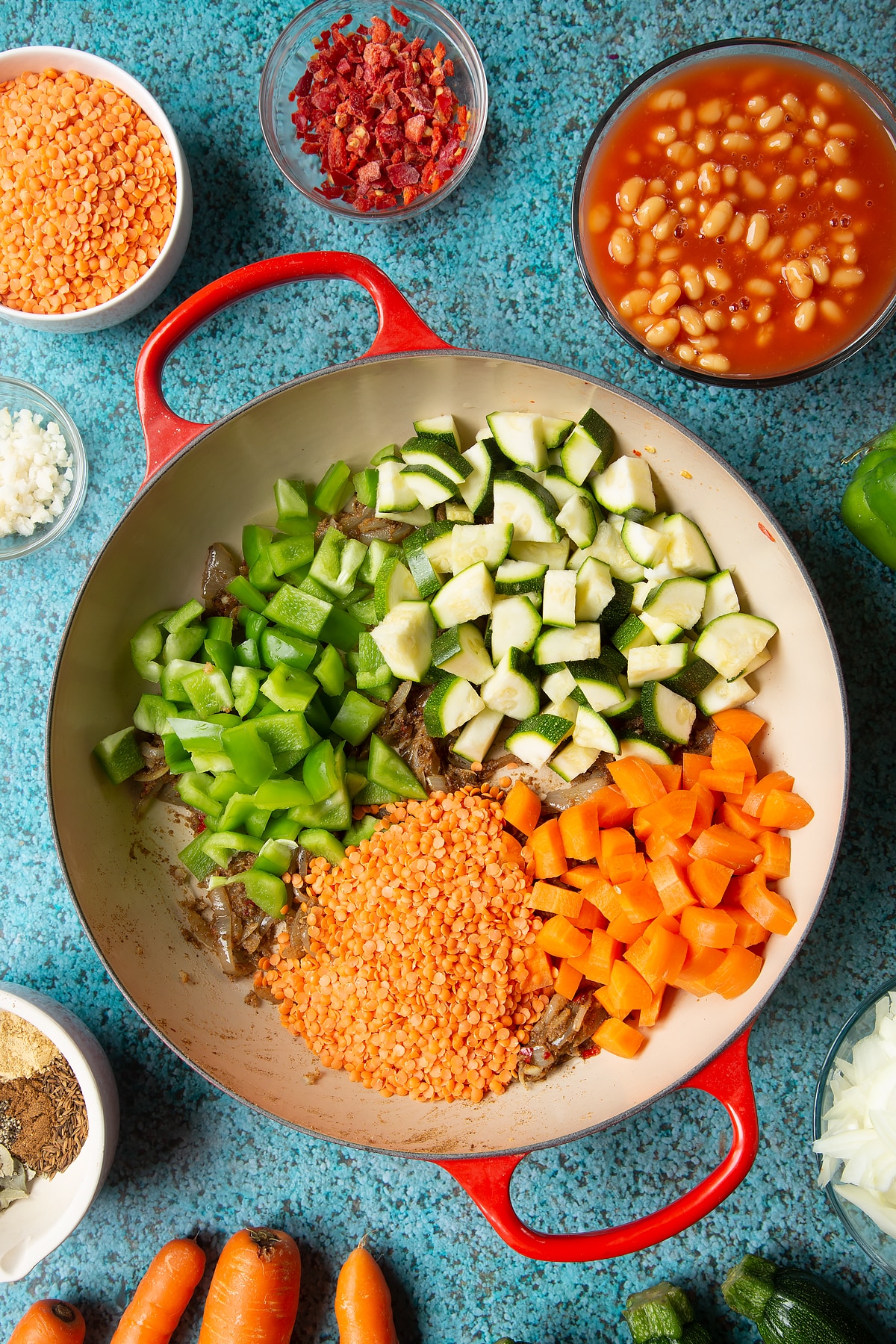Fried onions and spices in a pan with chopped peppers, carrots, courgette and red lentils. Ingredients to make baked bean curry surround the pen.