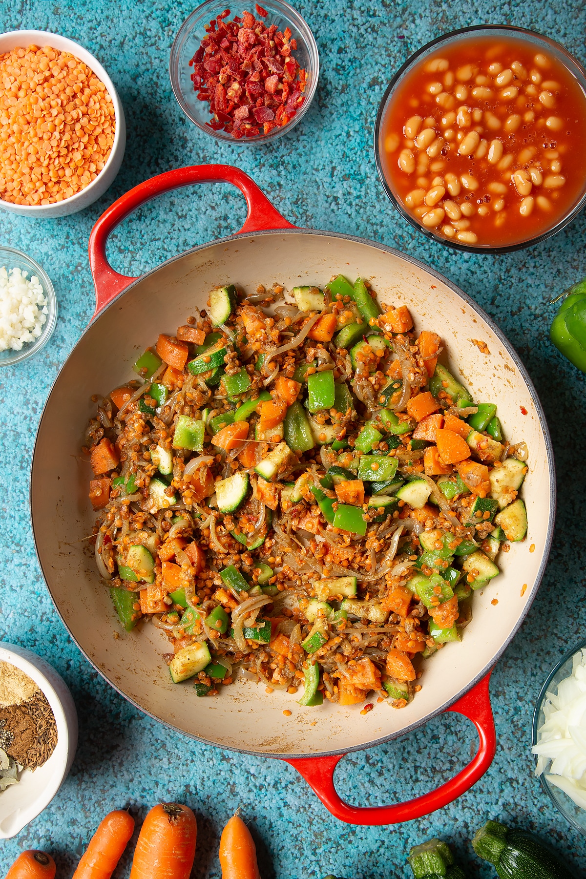 Fried onions, spices, peppers, carrots, courgette and red lentils in a pan. Ingredients to make baked bean curry surround the pen.