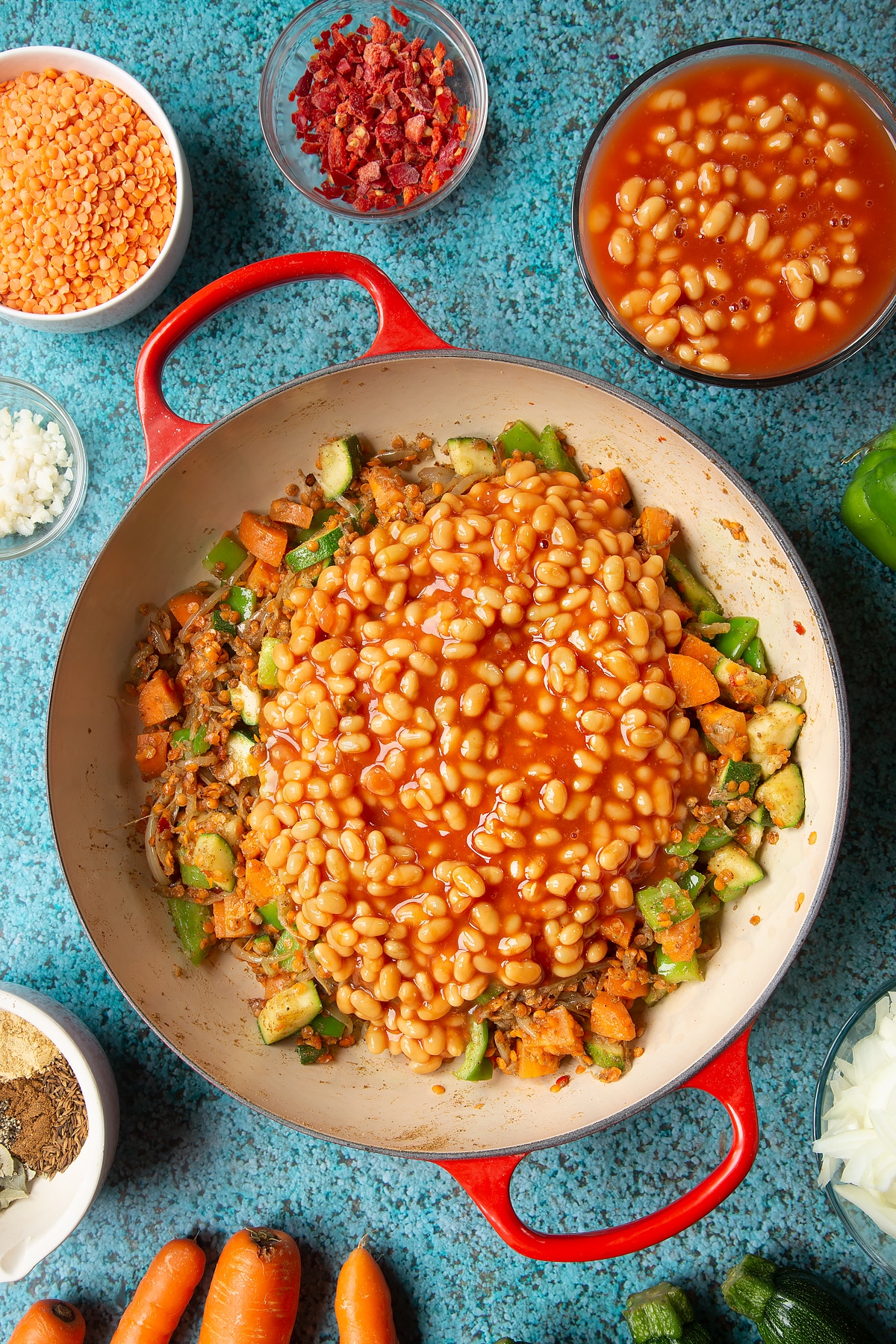 Fried onions, spices, peppers, carrots, courgette and red lentils in a pan with baked beans on top. Ingredients to make baked bean curry surround the pen.
