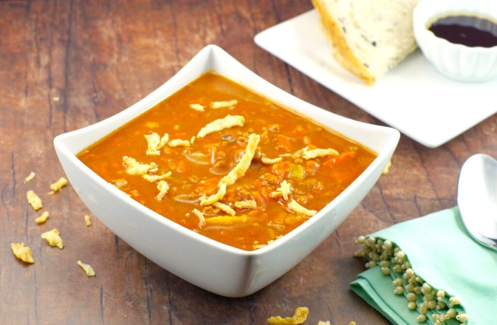 A square bowl of Creamy Carrot Soup on a dark brown wooden tabletop. At the side is a mint green napkin and a plate of bread with dip.