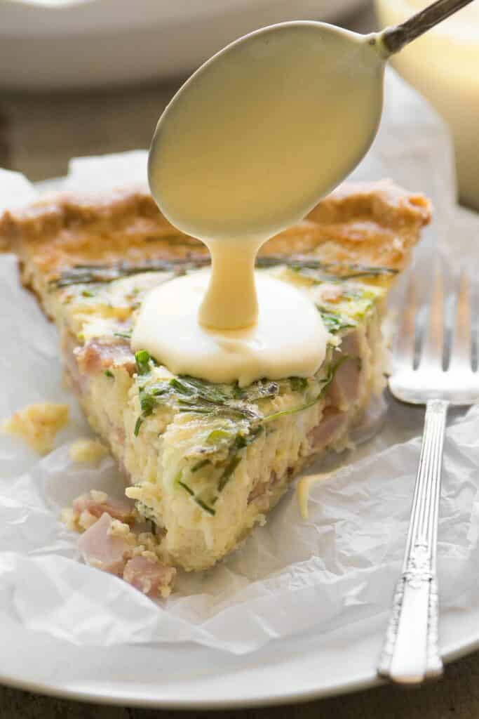 A slice of Eggs Benedict Quiche being drizzled in hollandaise sauce. A deocrative fork sits at the side.