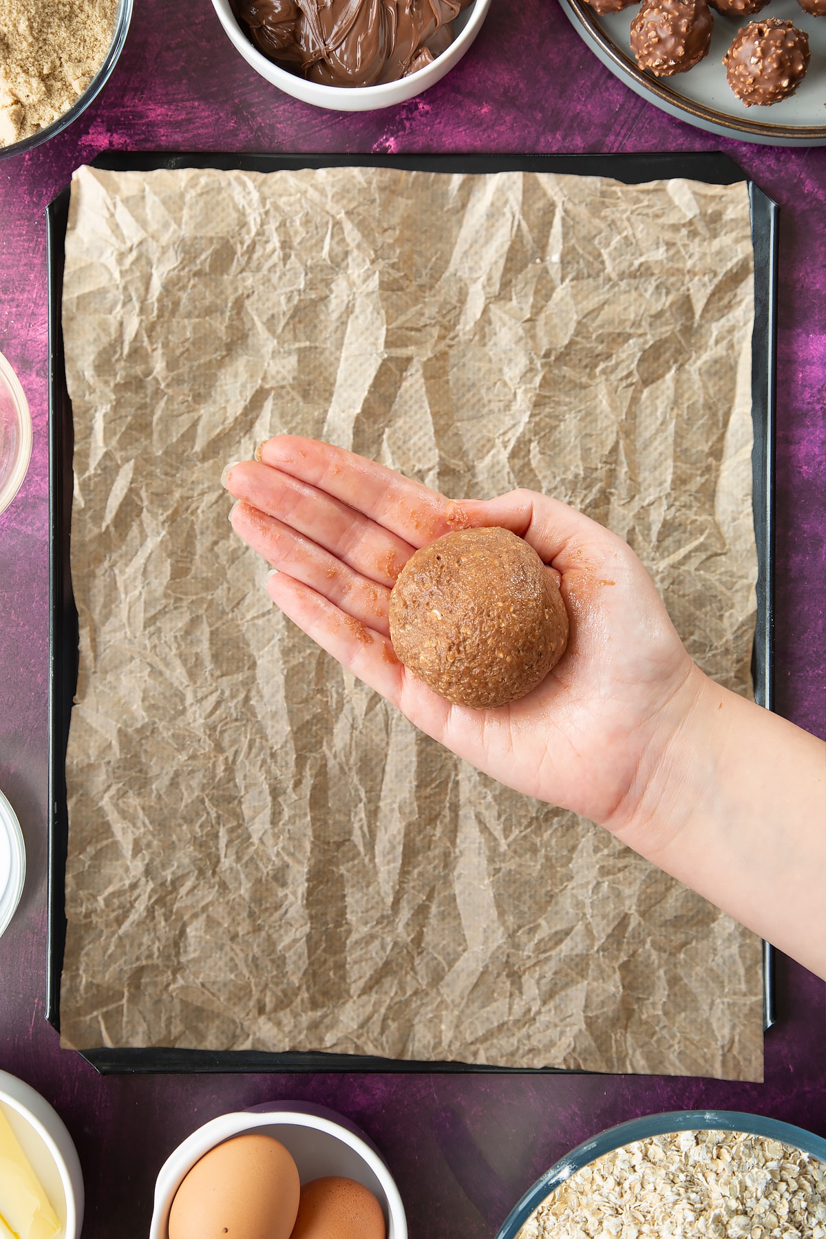 A hand holding a ball of oaty chocolate cookie dough. Ingredients to make Ferrero Rocher cookies surround a tray below.