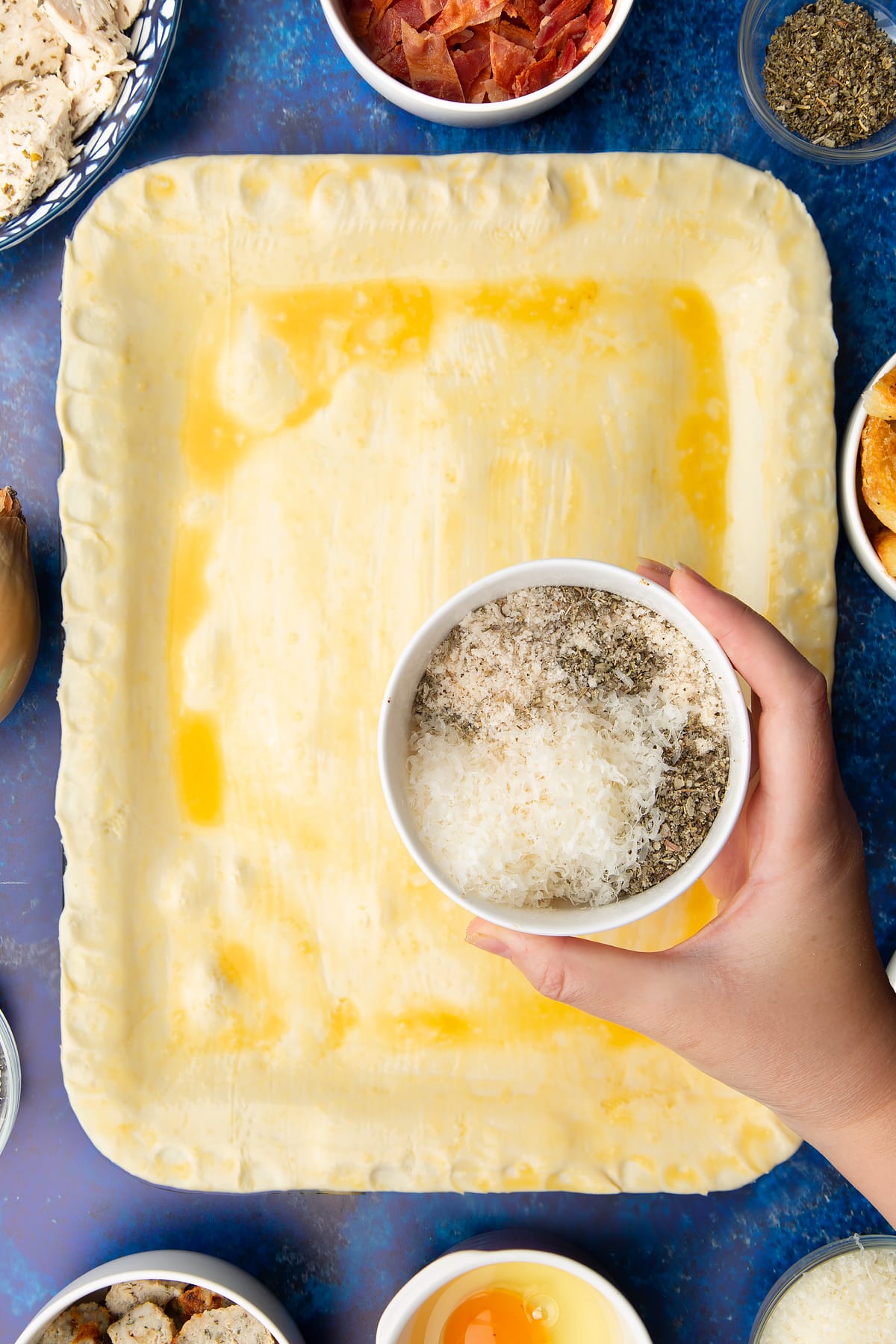 A roasting dish topped with a raw puff pastry lid that has been brushed with egg. Above the pie, a hand holds a bowl with breadcrumbs, cheese and herbs.