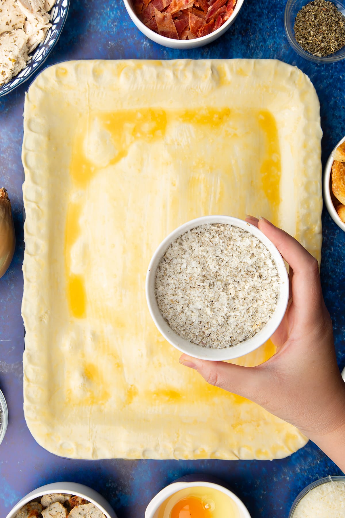 A roasting dish topped with a raw puff pastry lid that has been brushed with egg. Above the pie, a hand holds a bowl with breadcrumbs, cheese and herbs mixed together.
