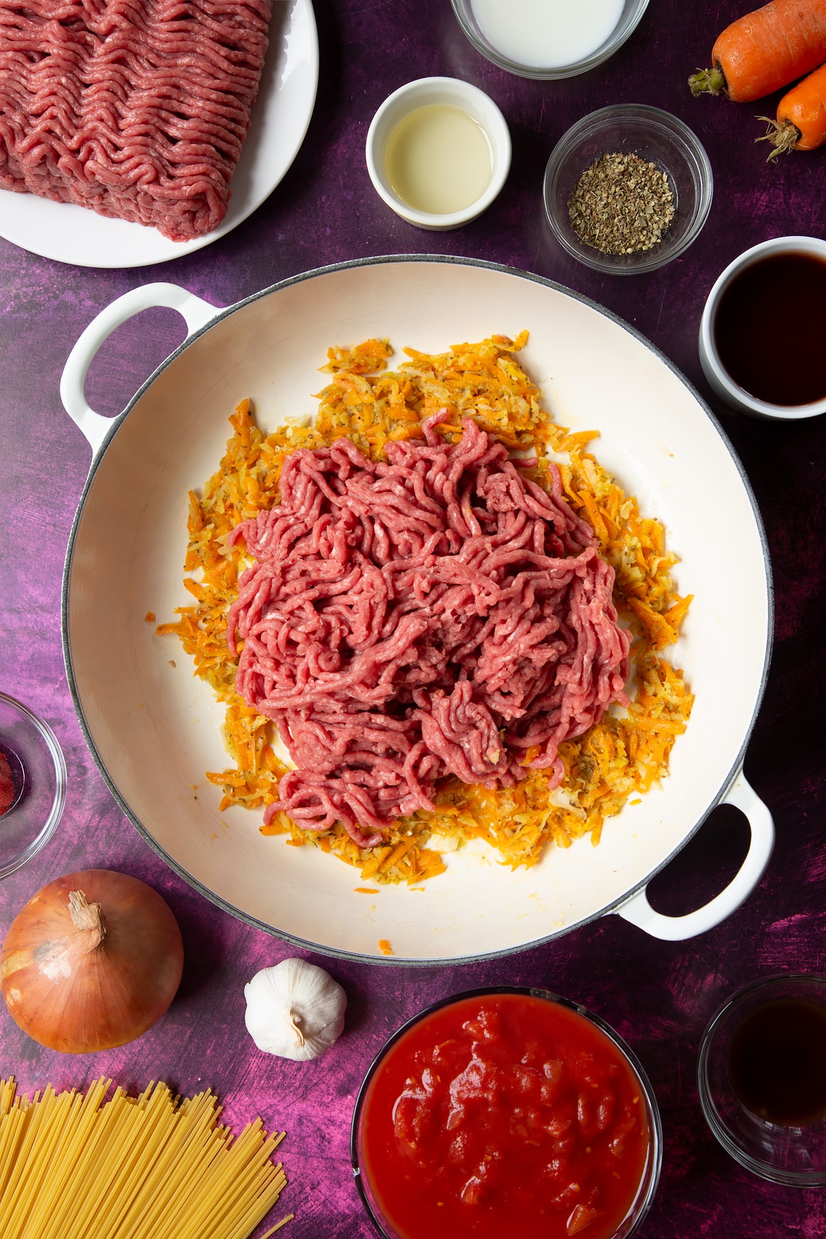 A large frying pan with fried carrot, onion, garlic and dried oregano. Raw lean beef mince has been added. Ingredients to make spaghetti bolognese Gordon Ramsay style surround the pan.