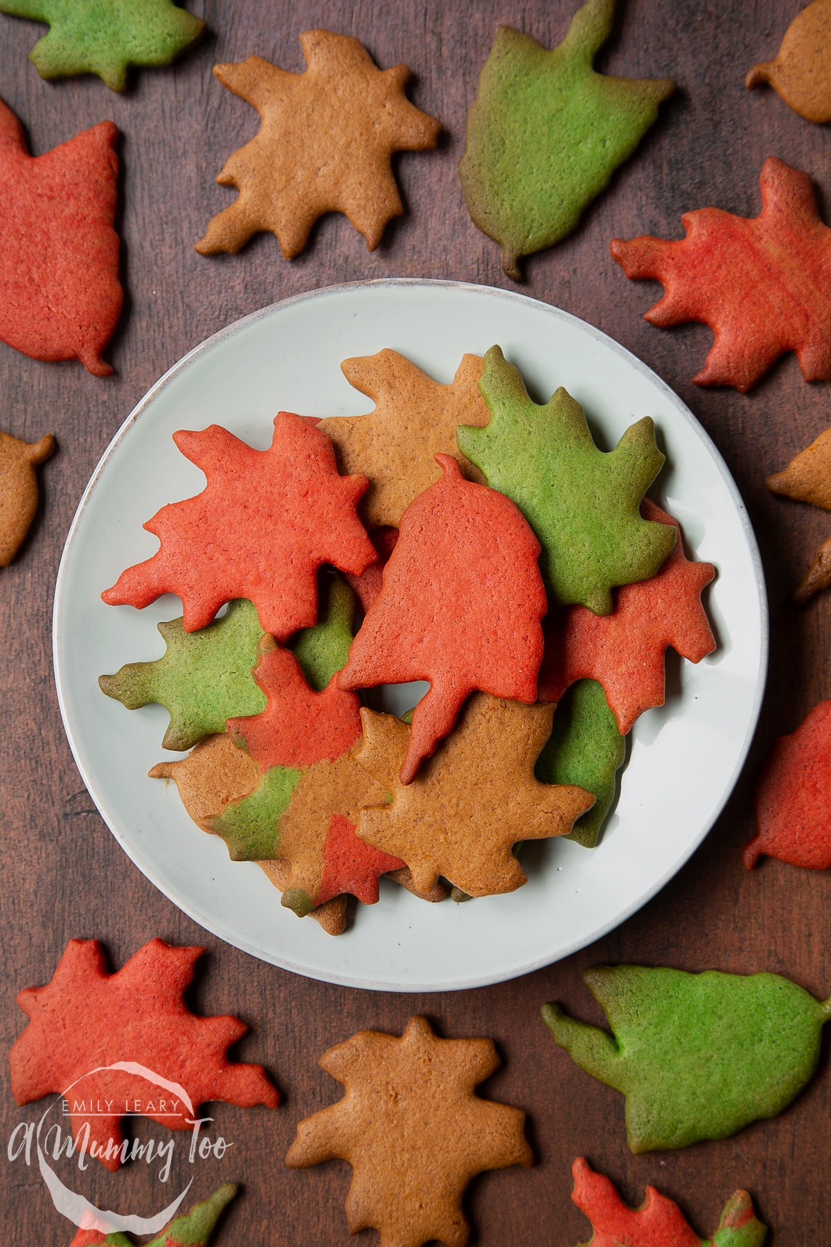 A plate of colourful red, green and brown autumn cookies cut into the shapes of autumn leaves. A red leaf biscuit sits in the centre.