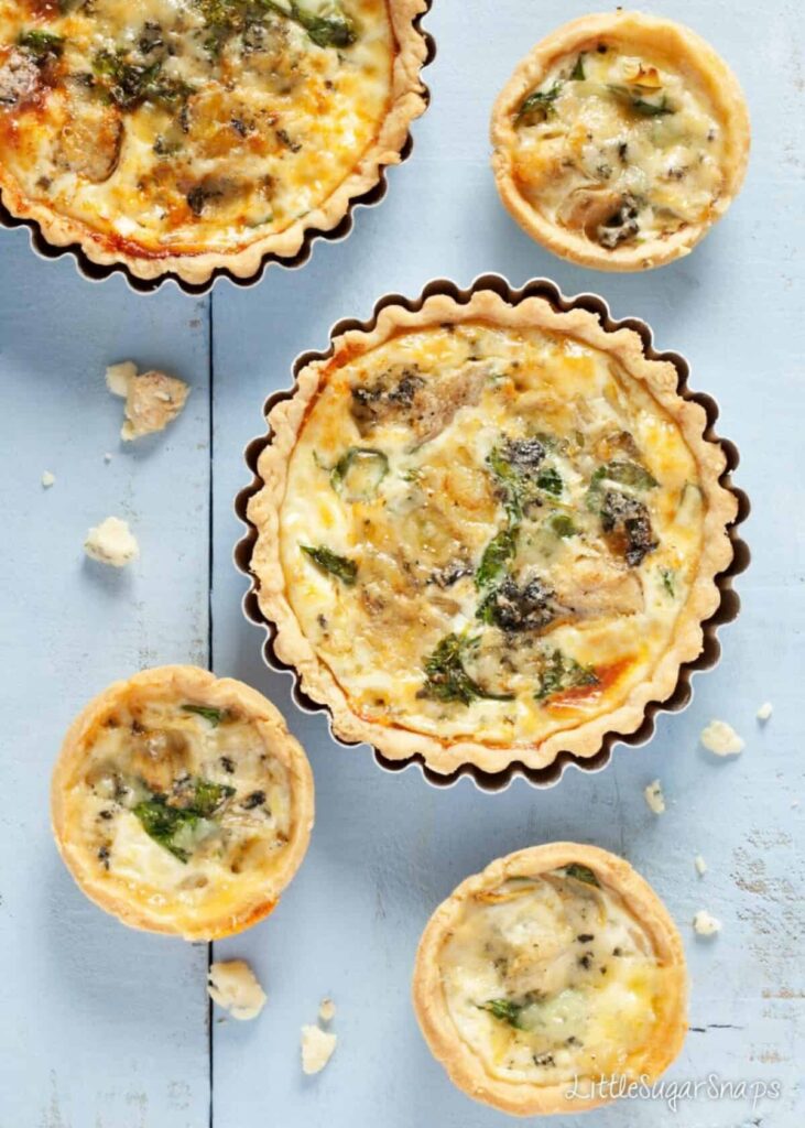 Five blue cheese, watercress and artichoke tarts sit on a white background. Three are smaller and two are larger and sit inside a quiche baking tray.
