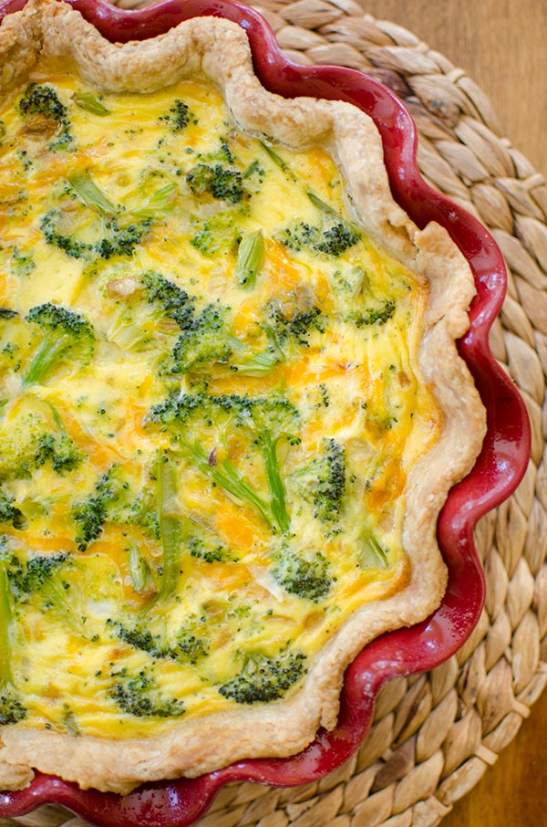 Broccoli Cheddar Quiche inside a red decorative dish on top of a wicker placemat. The quiche image is positioned on the left hand side and is cut off.