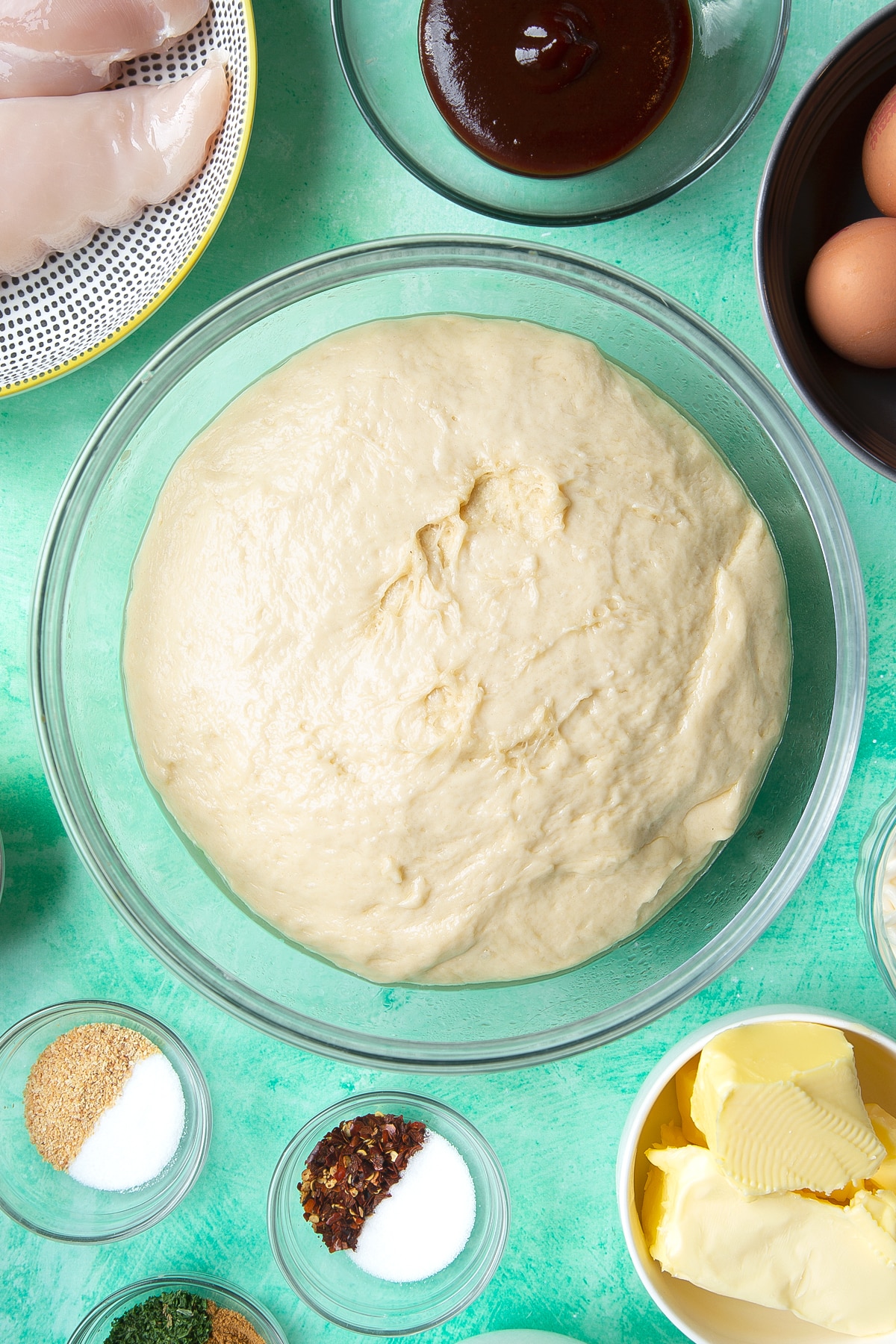 Proved bread dough ball in a glass mixing bowl. Ingredients to make chicken doughnuts surround the bowl.
