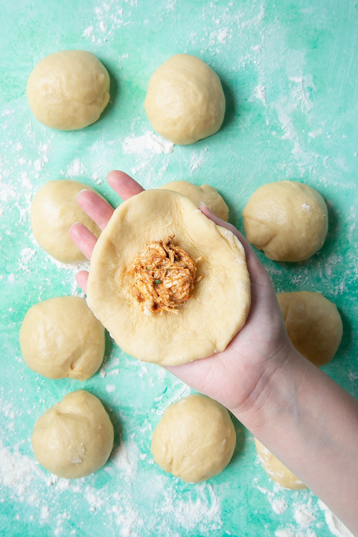 A hand holding a flatten round of bread dough about 12cm (4 3/4 inches) diameter. A spoonful of pulled barbecue chicken sits in the centre.