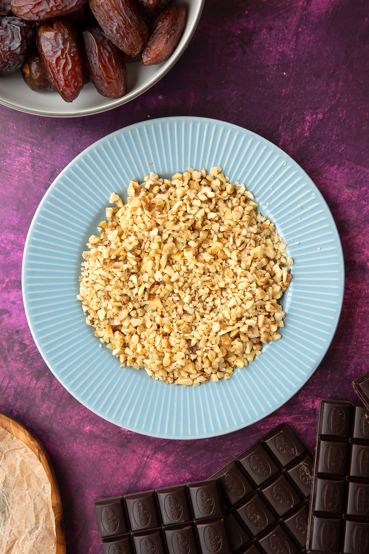Chopped walnuts on a blue plate, surrounded by ingredients to make chocolate dates.