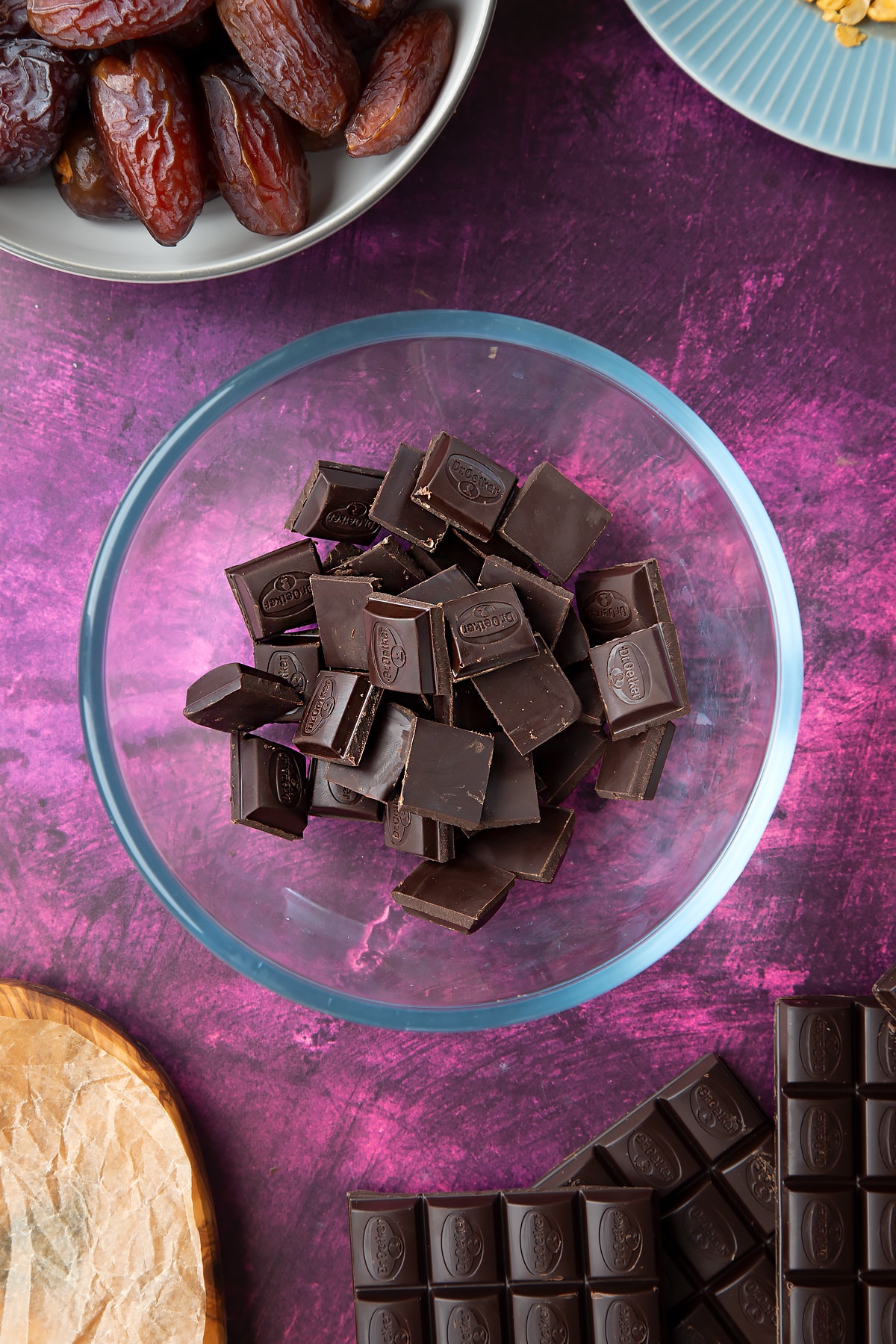 Dark chocolate squares in a bowl, surrounded by ingredients to make chocolate dates.