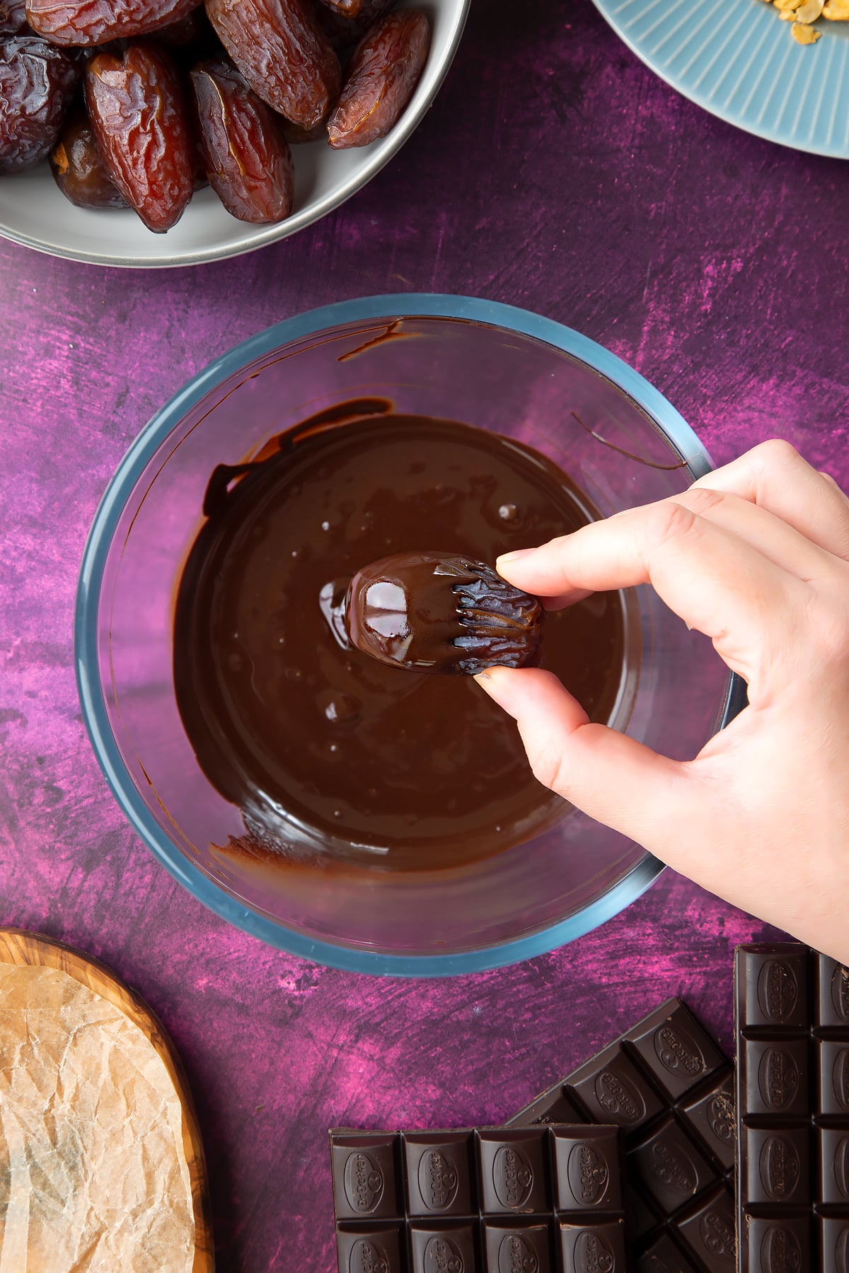 Melted chocolate in a bowl, surrounded by ingredients to make chocolate dates. A hand holds a date above the bowl, which is half coated with chocolate.