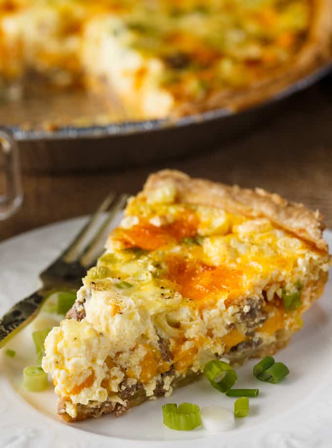 A Hamburger Quiche slice on a white plate with a fork at the side. The quiche slice is decorated with finely chopped chives. In the background you can see the full quiche sitting in a pie tin.