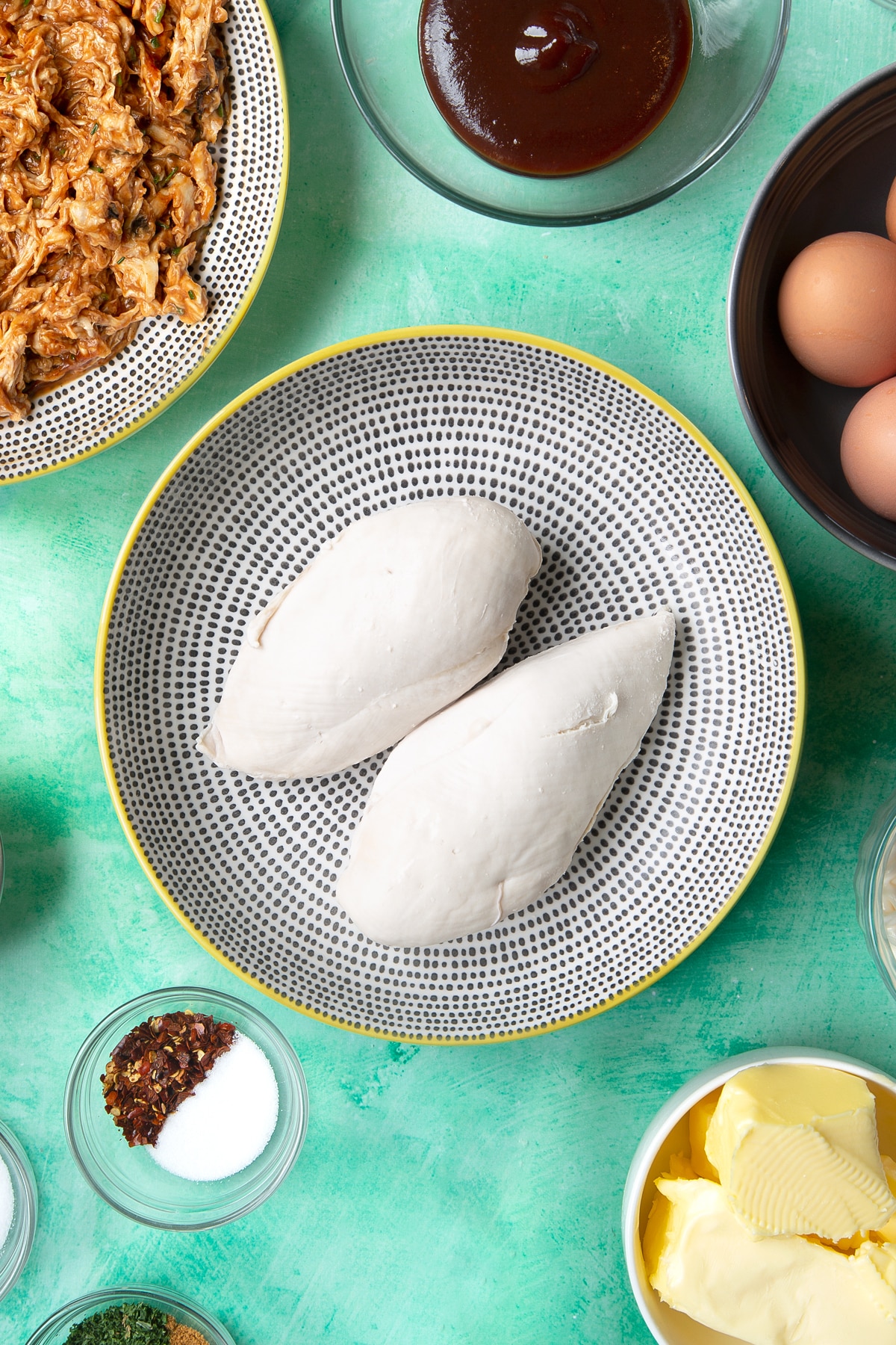 Two cooked chicken breasts in a pan. Ingredients to make chicken doughnuts surround the bowl.