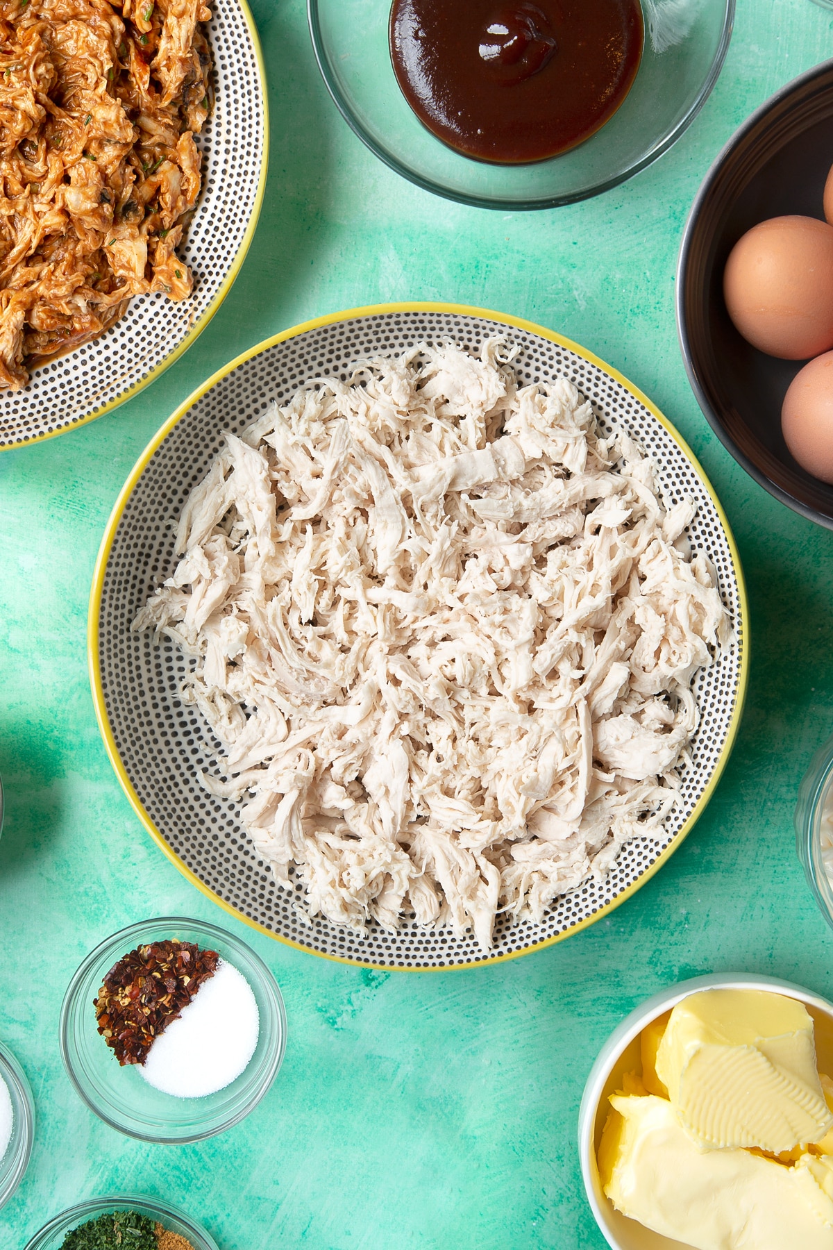 Shredded chicken breast in a bowl. Ingredients to make chicken doughnuts surround the bowl.