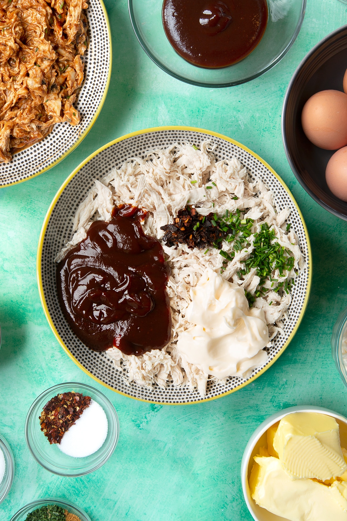 Shredded chicken breast in a bowl, topped with barbecue sauce, mayo, chilli flakes and chopped chives. Ingredients to make chicken doughnuts surround the bowl.