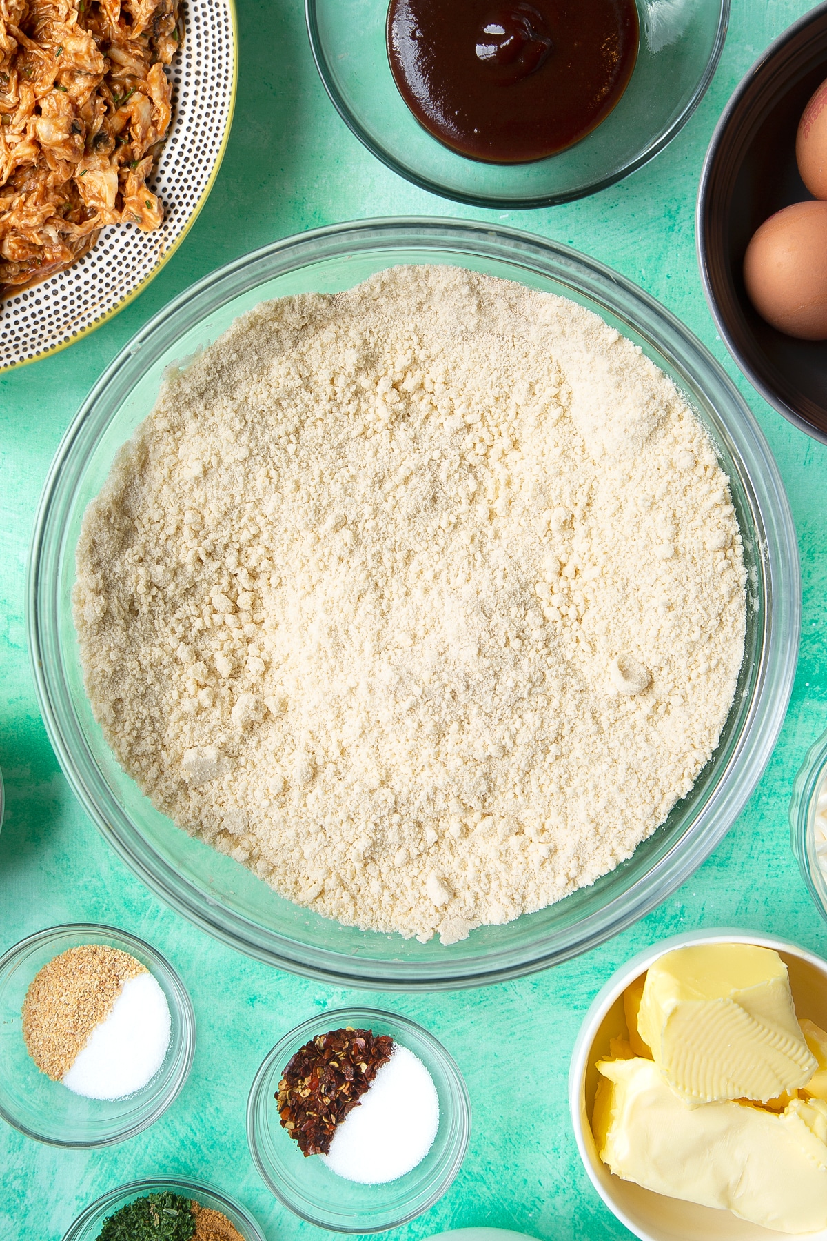 Flour and butter rubbed together with salt, sugar and yeast in a glass mixing bowl. Ingredients to make chicken doughnuts surround the bowl.