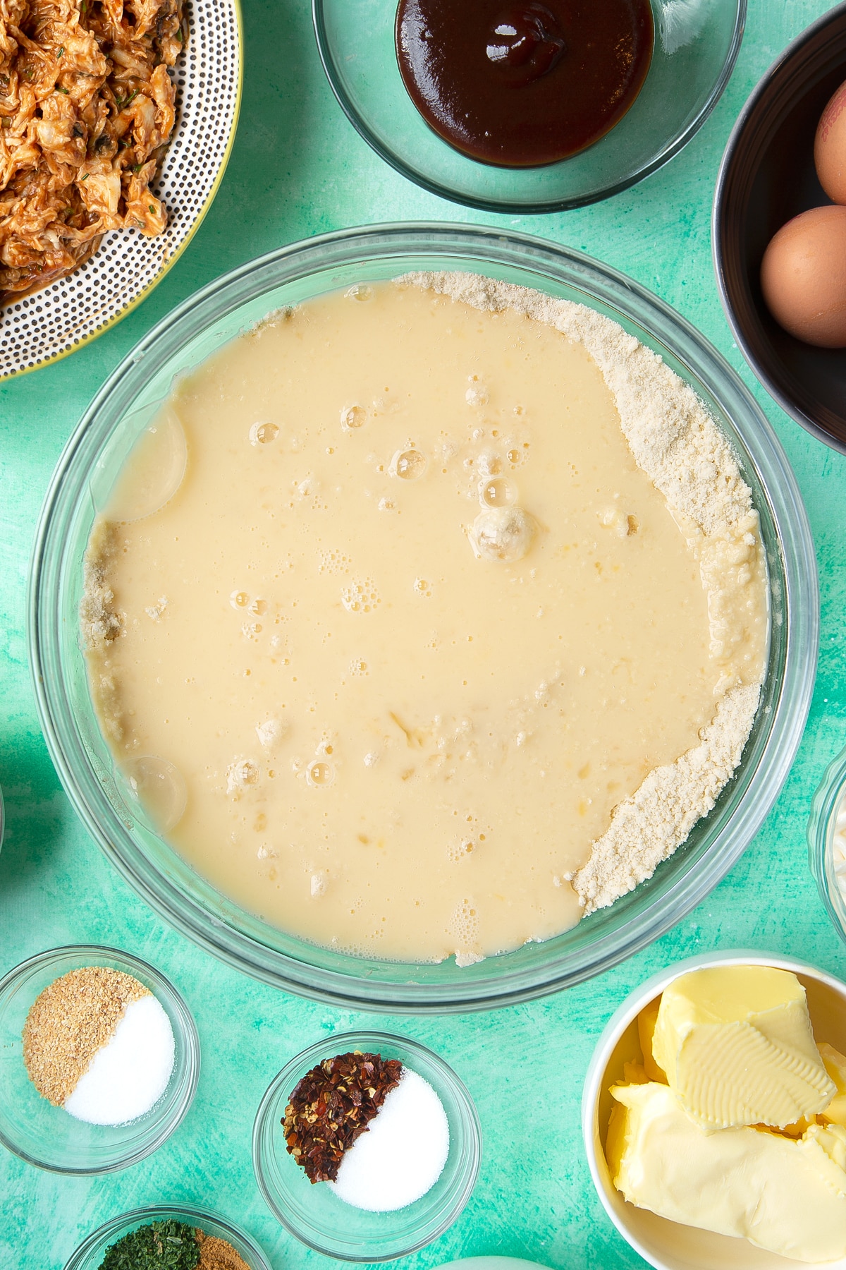 Flour and butter rubbed together with salt, sugar, yeast eggs and water in a glass mixing bowl. Ingredients to make chicken doughnuts surround the bowl.