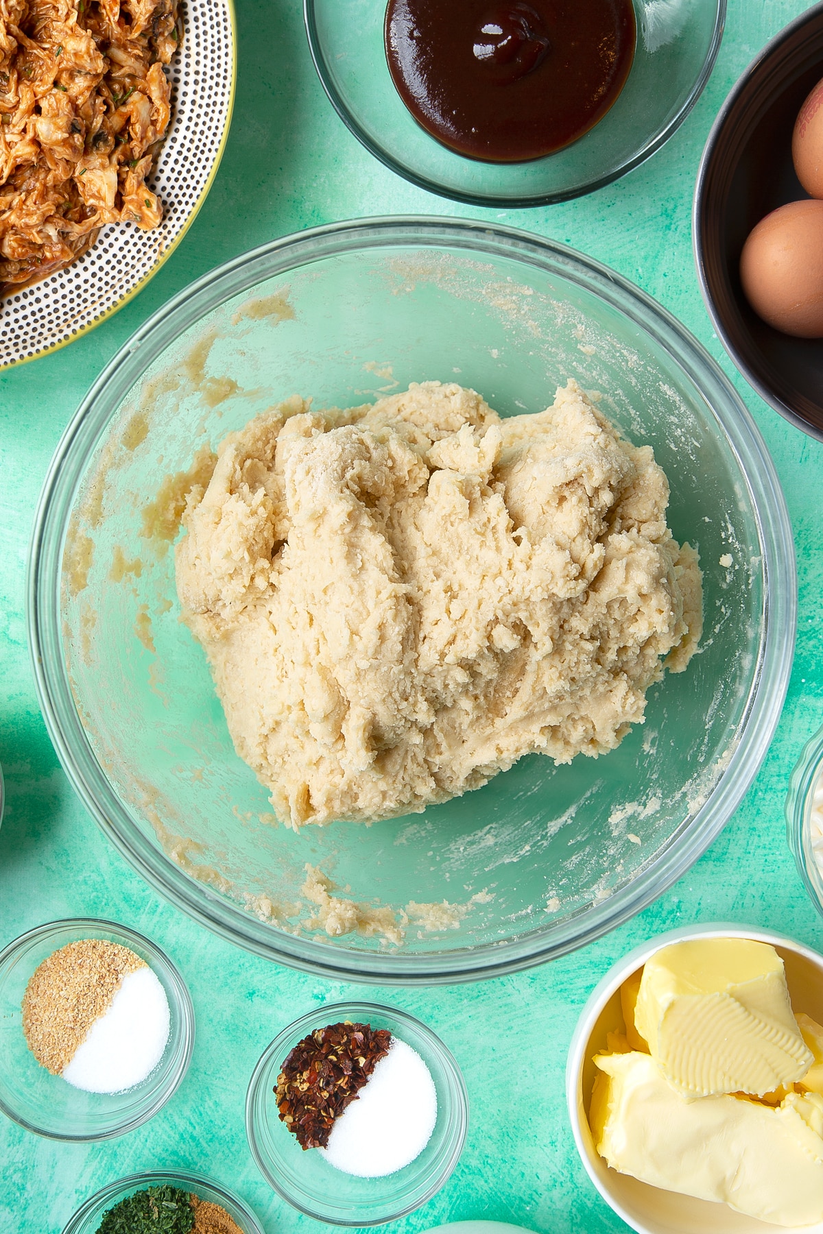 A roughly mixed bread dough in a glass mixing bowl. Ingredients to make chicken doughnuts surround the bowl.