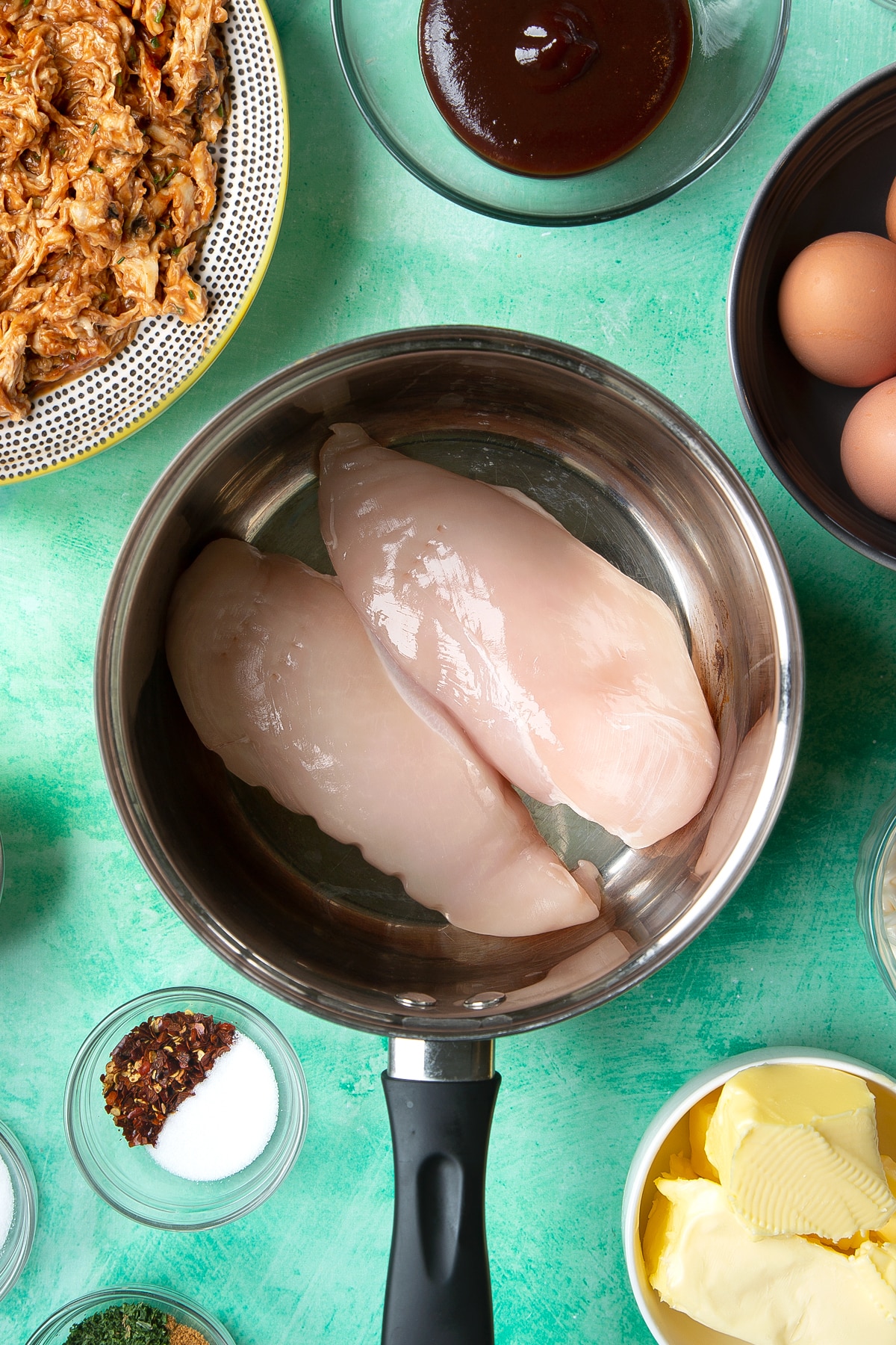 Two raw chicken breasts in a pan. Ingredients to make chicken doughnuts surround the pan.