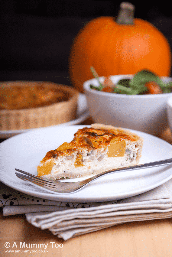 Roast Pumpkin & Caramelised Onion Quiche slice sits on a white plate in the foreground with a fork at the side. The plate sits on a handful of napkins. In the background there's a bowl of salad and a whole pumpkin.