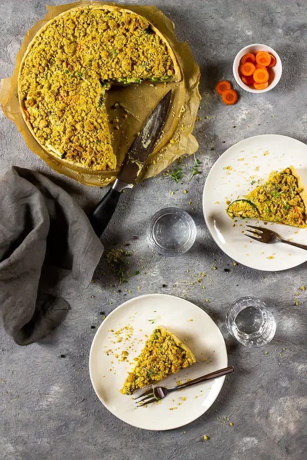 A large Garden Veggie Quiche With Cheesy Crumbs with two slices cut out. Both of the slices are situated on a white plate with a fork at the side.
