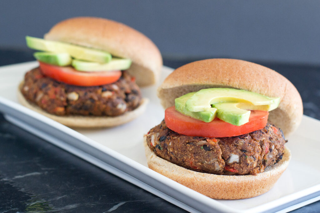 Two black bean burgers with buns, avocado slices and tomatoes on a white rectangular plate.