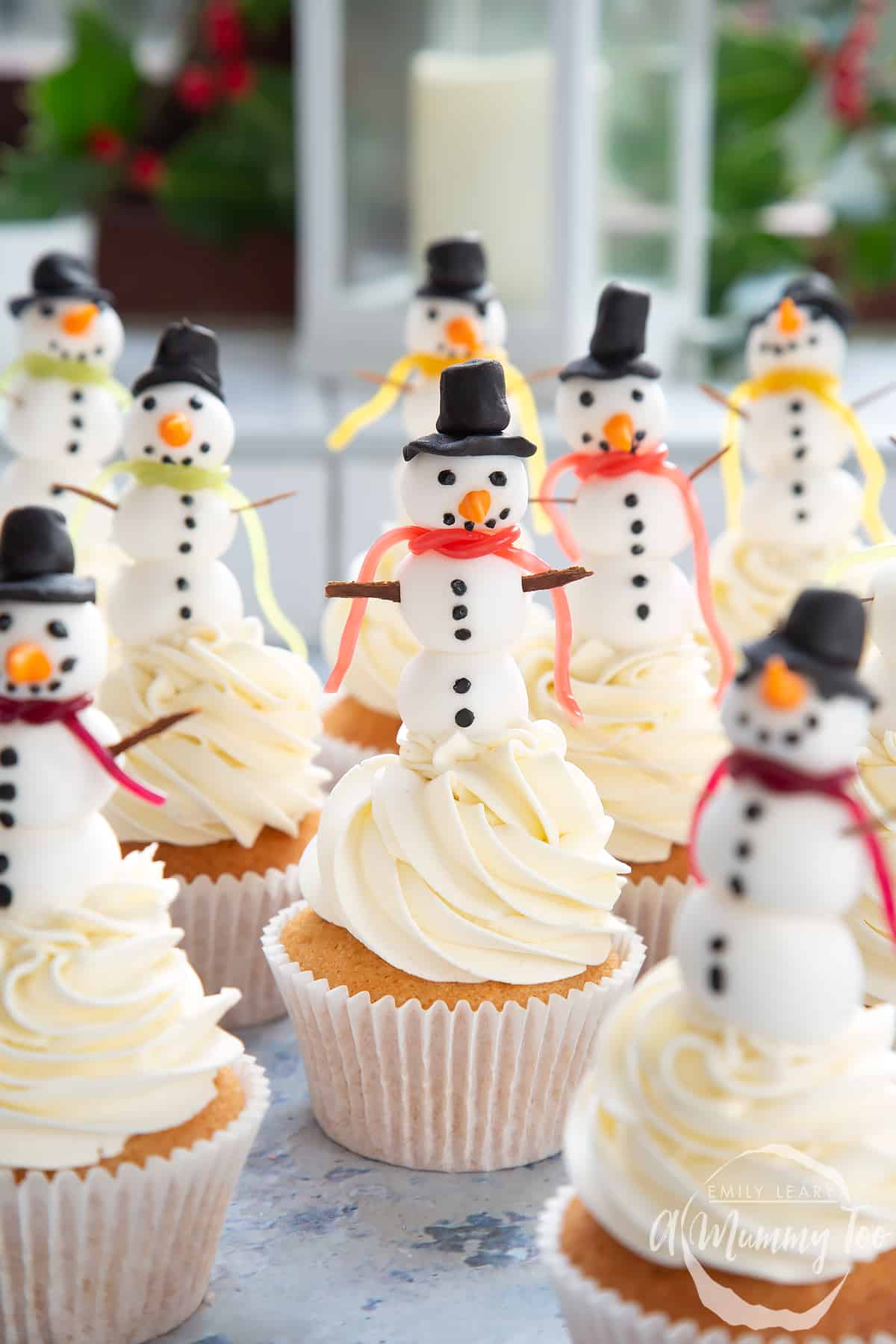 Multiple snowman cupcakes sat on a table with festive decorations in the background.