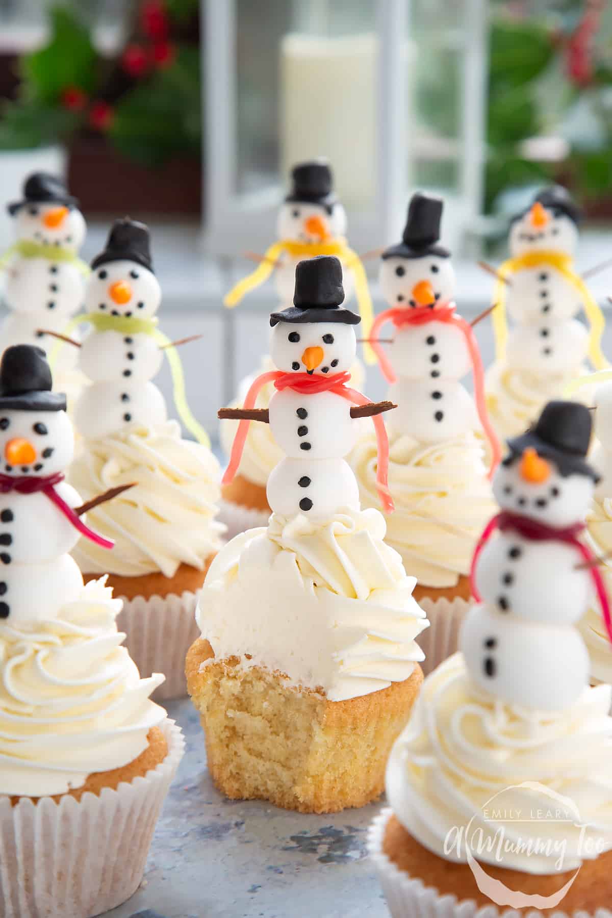 Multiple snowman cupcakes on a table with a festive background. The centered snowman cupcake has been unwrapped and a bite taken out.