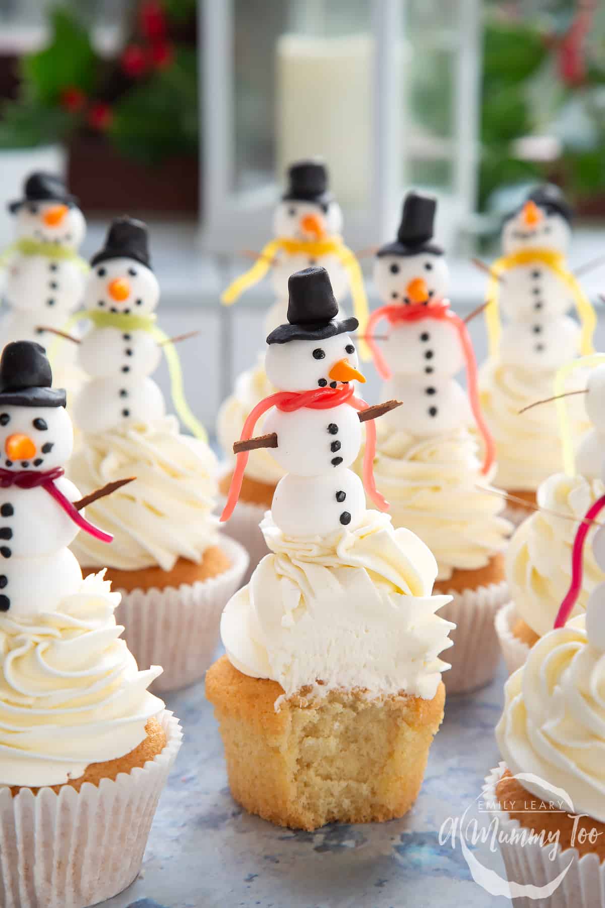 Multiple snowman cupcakes sat on a table. The centered snowman cupcake has been upwrapped and a bite has been taken out.