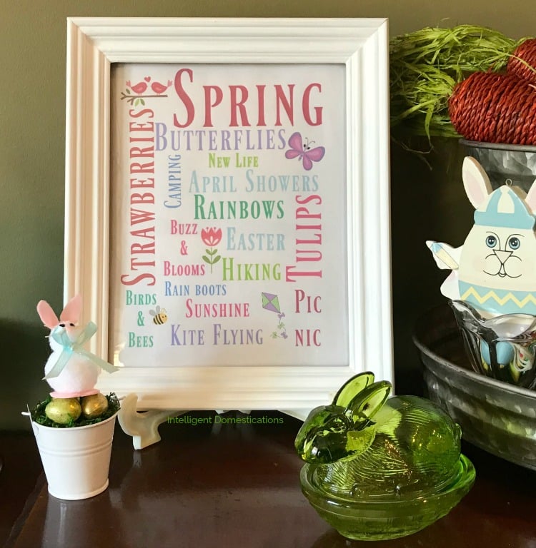 A framed artwork on an Easter decorated shelf. The art is a series of spring-themed words in coloured fonts.