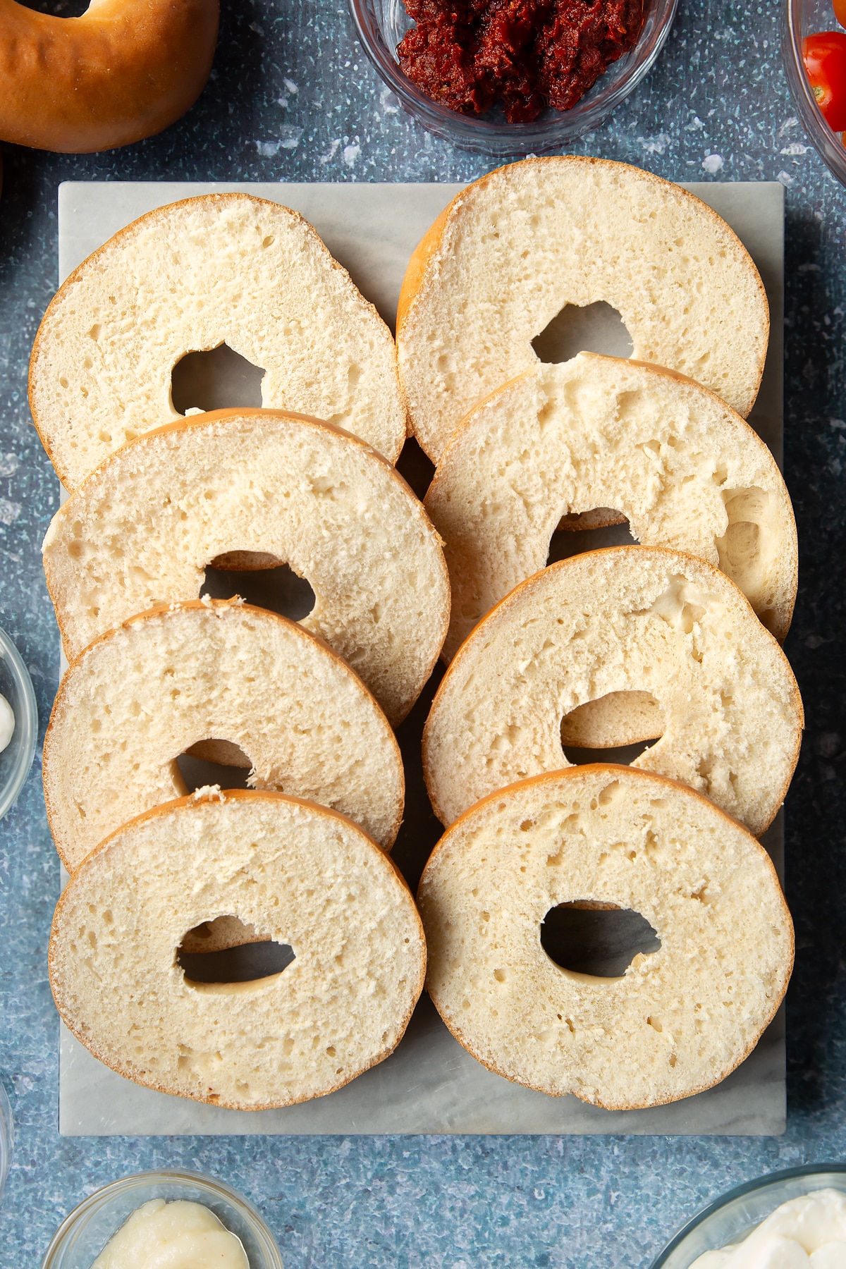 Sliced open bagels on a marble board. Ingredients to make halloumi bagels surround the board.