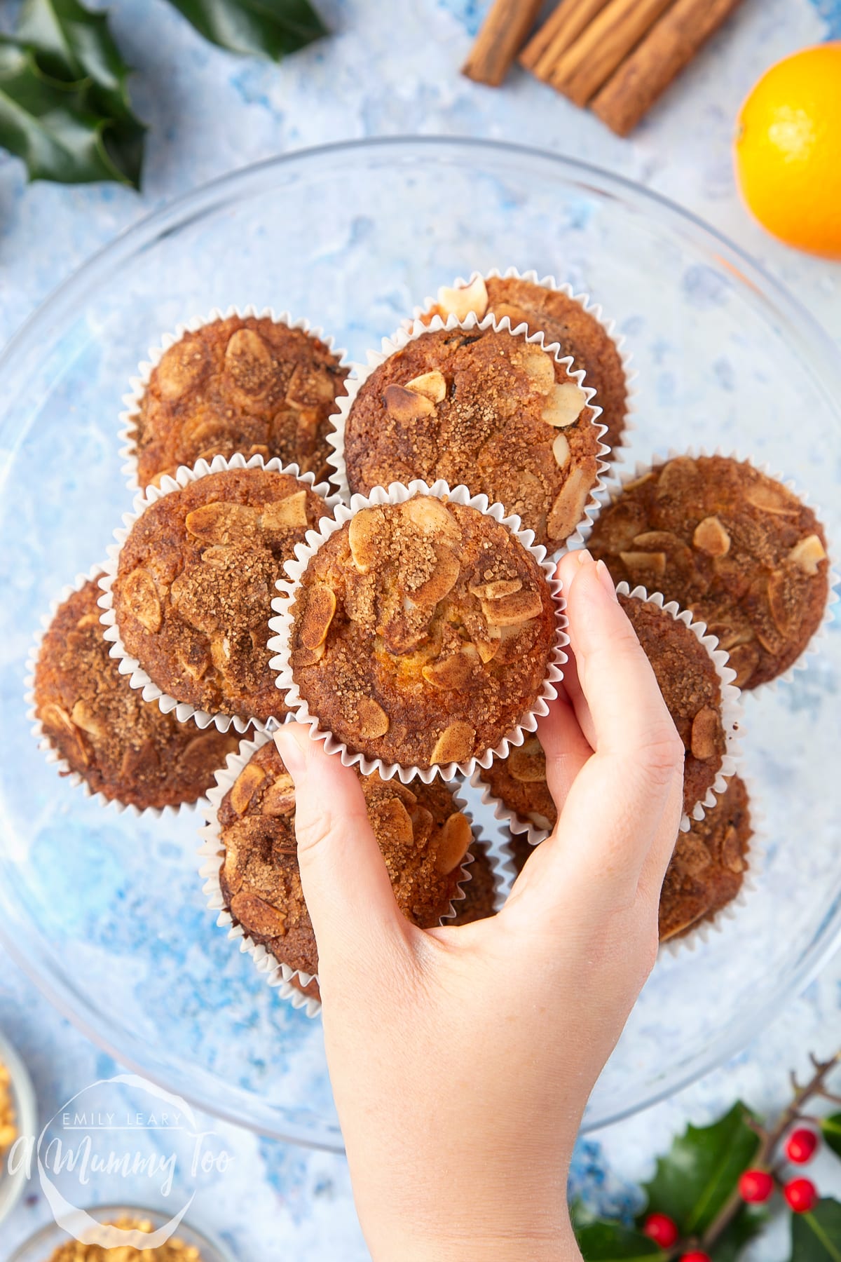 Overhead shot of a hand touching a Orange and cinnamon muffin with a mummy too logo in the lower-left corner